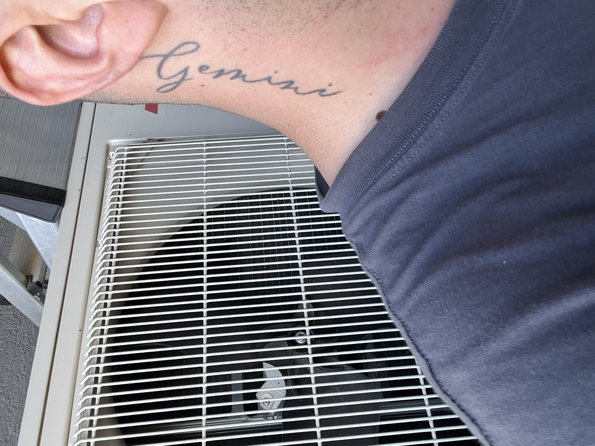 A close-up shot shows the side of a person's neck with a tattoo of the word "Gemini" in cursive script. The person is wearing a dark grey t-shirt. Behind them is the grill of an outdoor air conditioning unit. The background appears to be an exterior wall of a building. The scene is outdoors in daylight. The location is Varna, Bulgaria.