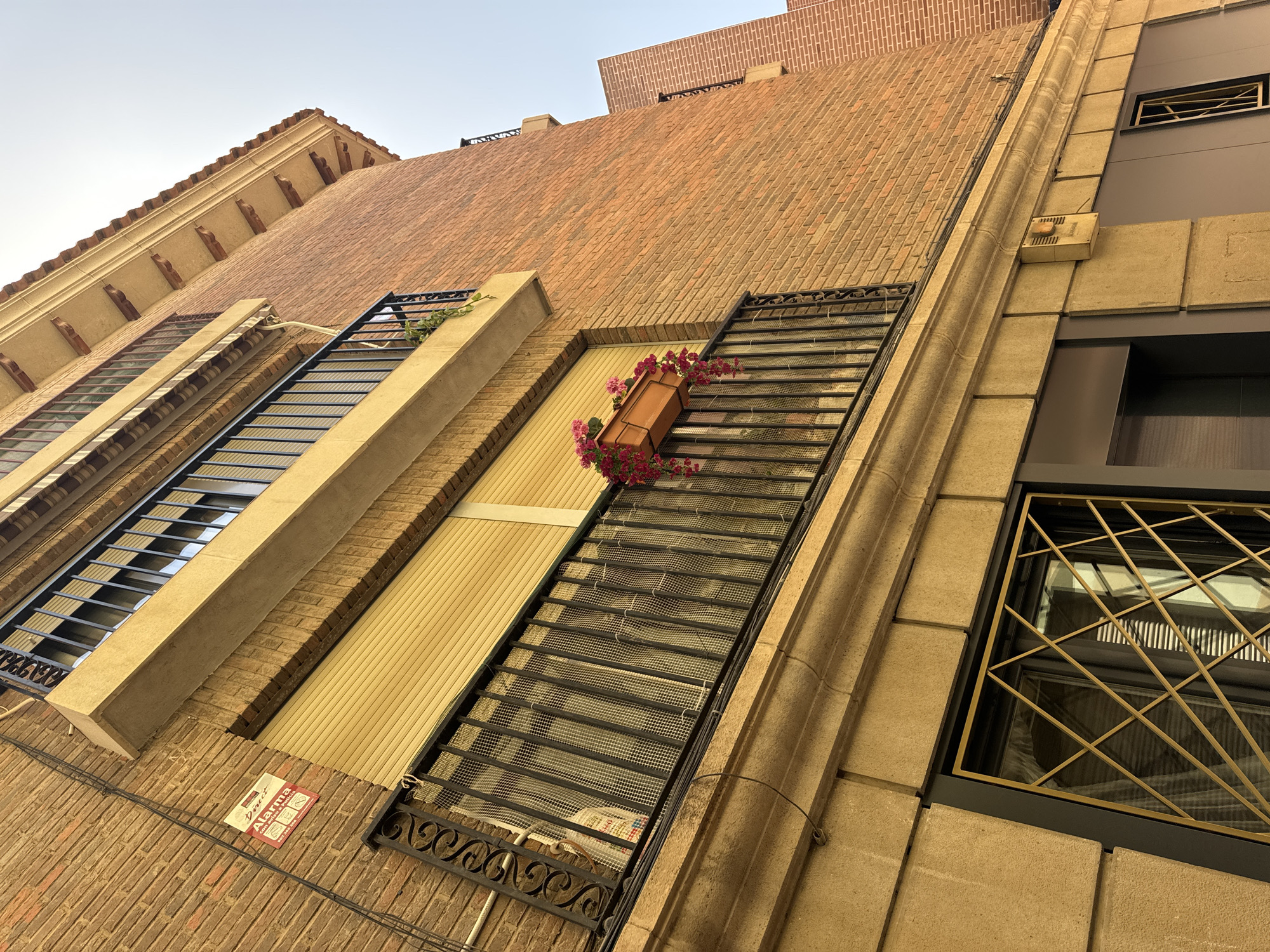 This is a low-angle, full shot of an urban building facade, taken on a clear day. The building has a multi-layered design, with a terracotta tiled roof visible at the top. The facade itself is constructed from brick and stone.
There are several windows, many of which are protected by metal grilles. One prominent window features a planter box with pink flowers. Another window, to the right, has an intricate, diamond-shaped gold grille. Various architectural details, such as decorative cornices and a vertical stone element with carved lines, are visible.
On the brickwork of the lower section of the building, a red and white sign is attached. The text on the sign is partially visible and appears to be an alarm company advertisement, with the word "Alarma" clearly legible. There are also electrical wires running along the facade. The scene depicts a quiet urban setting, with no people visible. The time of day is likely daytime, given the bright, clear sky.