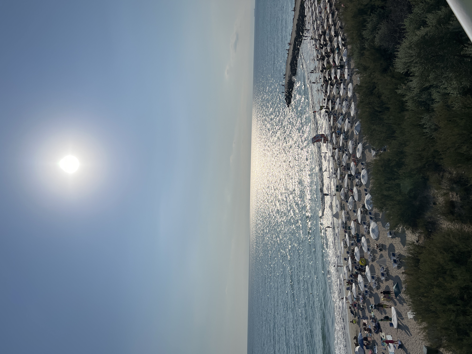 This is a high-angle, outdoor, eye-level shot of a crowded beach in Pomorie, Bulgaria, on a sunny day. The sky is a clear, bright blue with the sun prominently featured in the upper center of the frame, creating a strong glare on the sea. The sea itself is a deep blue, with waves lapping at the shore. The beach is covered in a dense arrangement of white beach umbrellas and lounge chairs, with many people visible relaxing, sitting, and walking. In the foreground, dense green bushes and trees line the edge of the beach. A stone pier extends into the sea from the right side of the frame, with several people standing on it. The water is glittering with sunlight, and a faint, hazy line of the horizon is visible in the distance.