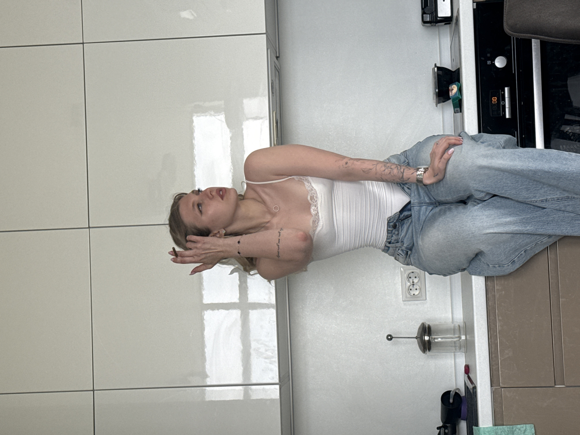 A woman is posing in what appears to be a kitchen. She is wearing a white tank top and light blue jeans, and she has tattoos on her arms. She has light skin and blonde hair. She is wearing a watch on her left wrist. The kitchen has white cabinets and countertops. There is a built-in oven to the right of the woman. There is an electrical outlet and a kitchen appliance on the countertop near the woman. It is not possible to determine if this photo was taken in Rostov-on-Don, Russia.
