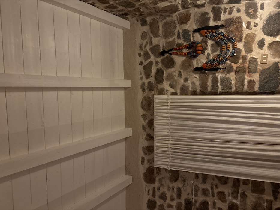 The interior corner shows a white, paneled wall meeting a stone wall. The stone wall has an art piece, possibly made of beads and fiber, shaped like a curved arrow. A window with closed, horizontal blinds is situated below the artwork. The room appears dim, emphasizing the textures and color differences of the materials.