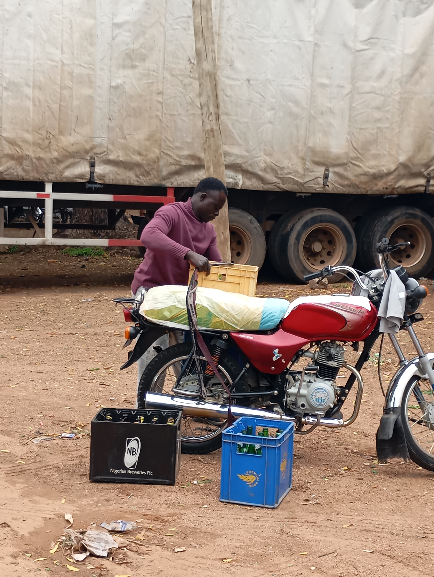 A Black male, wearing a maroon sweater and light-colored pants, is standing next to a red and white motorcycle. He is holding a yellow plastic crate filled with items, placing it on the back of the motorcycle. The motorcycle is parked on a dirt surface. To the left of the motorcycle, a black crate labeled "NB Nigerian Breweries Plc" is on the ground, containing bottles. To the right of the motorcycle, a blue plastic crate with bottles is also on the ground. In the background, a large truck with a beige tarp-covered trailer is visible, with its tires touching the ground. The scene appears to be outdoors, with dry vegetation and debris scattered on the ground. The overall environment suggests a roadside or loading area in Shendam, Nigeria.