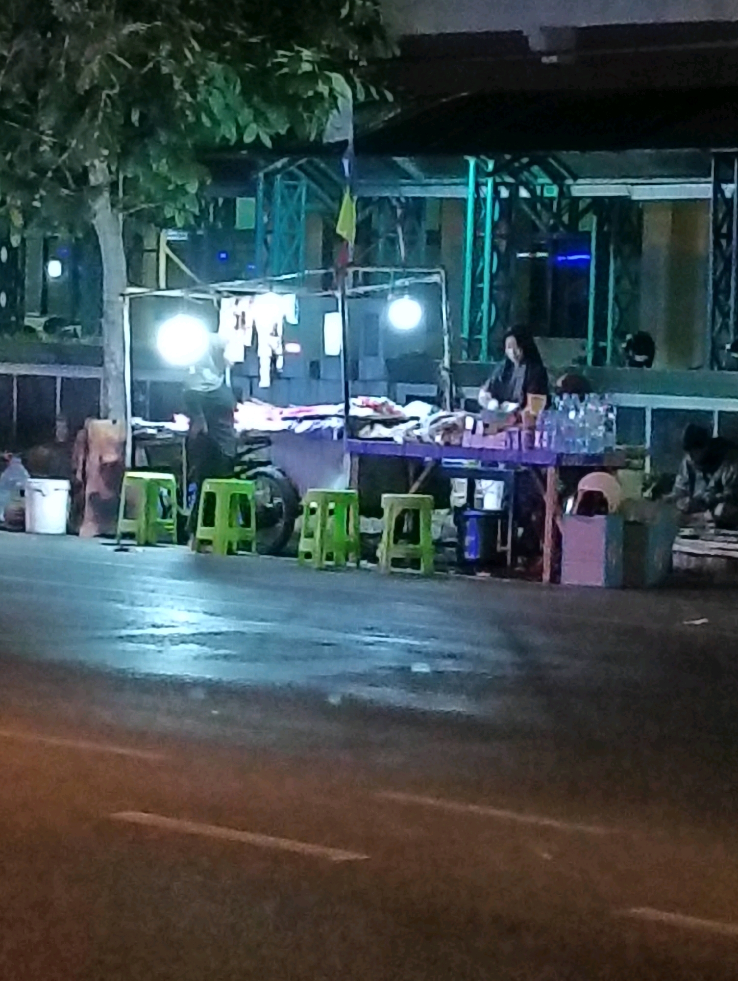The image depicts a street-level scene at night in Purworejo, Indonesia. The primary focus is a food stall or makeshift shop situated on a paved street, possibly a sidewalk or roadside. A vendor, a woman with dark hair wearing dark clothing, is visible behind a purple table laden with goods. She appears to be attending to something on the table. To her right, several bottles of liquid are stacked. In front of the stall, three bright green plastic stools are arranged on the ground. A motorcycle is parked behind the stall, with a bright light emanating from it, illuminating the area and casting a glare. To the left of the stall, there is a tree with dense foliage overhanging the scene, and a large white bucket is visible near a wooden object. In the background, structures with illuminated elements and what appears to be scaffolding or a metal framework are present. Further to the right, another person is seated on the ground. The street surface is wet, reflecting the ambient light. The overall impression is one of a bustling, albeit dimly lit, nighttime commercial activity in an urban setting.