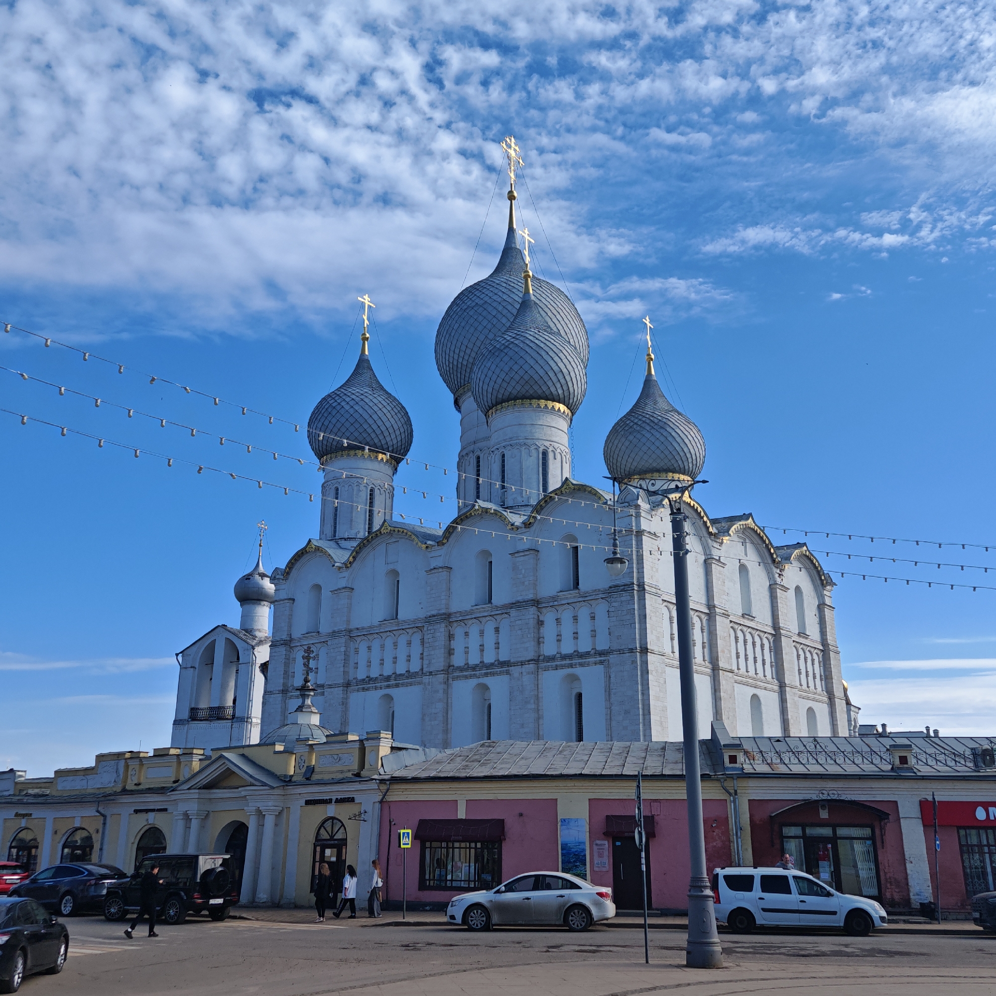 The image shows a large, pale stone building with distinctive bulbous domes topped with golden crosses against a bright blue sky with scattered clouds. Electrical lines with hanging lights run across the top of the frame. The building is set behind a row of lower, more colorful buildings painted in yellow, pink, and other hues. A few people are walking in front of the buildings on the sidewalk. Several cars are parked along the street in front of the buildings. This is likely a view of the Rostov Kremlin, located in Rostov Veliky, Russia.