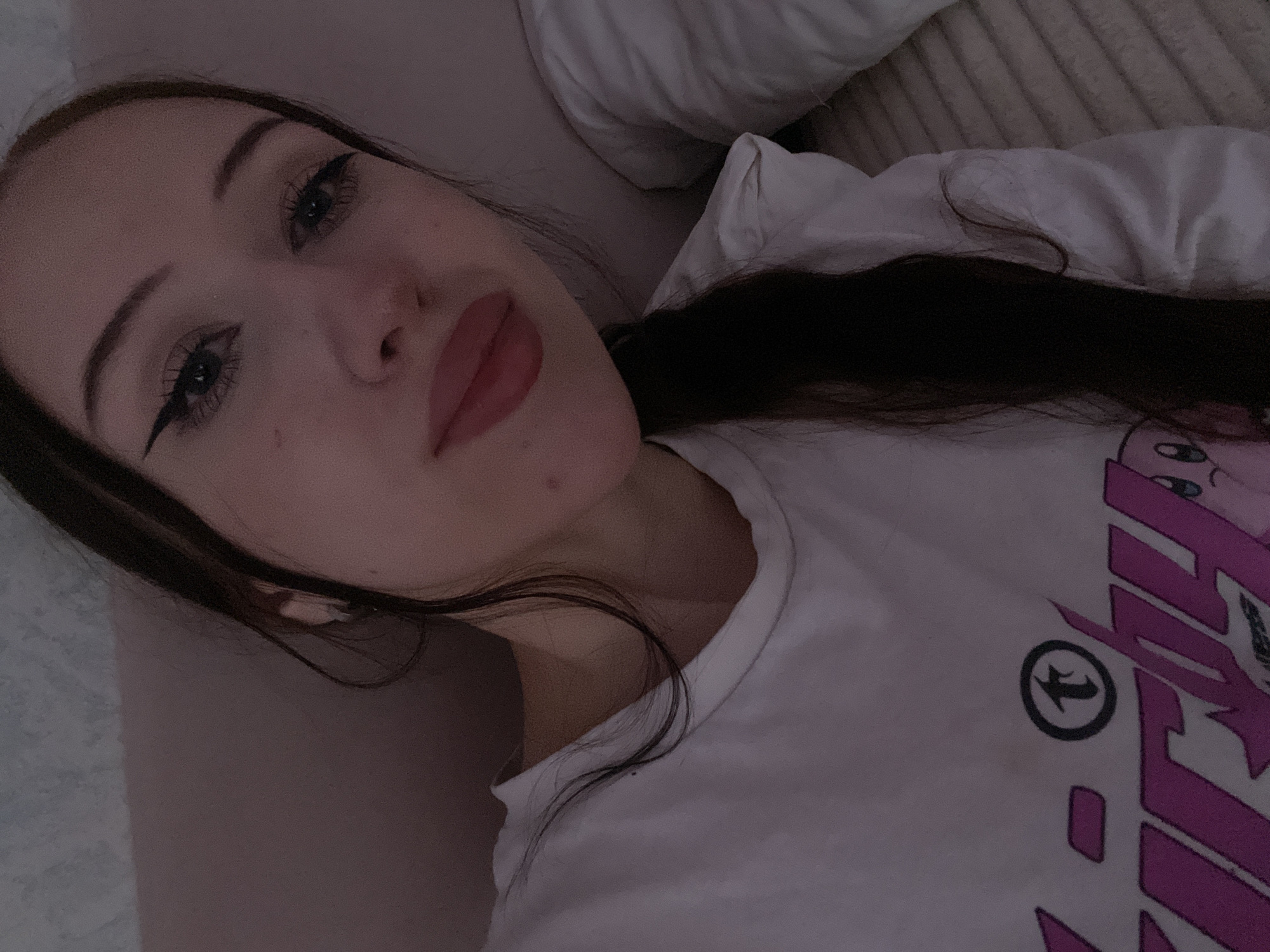 A young woman with long, dark hair and winged eyeliner is lying down and taking a selfie. She is wearing a white t-shirt with a pink graphic design. Her face is close to the camera, and she is looking directly at it. The background appears to be a bedroom, with a pillow visible on the right side of the image. The lighting is dim, suggesting it might be nighttime or a dimly lit room. There are no other people or objects clearly visible in the frame. The image was taken in Votkinsk, Russia.