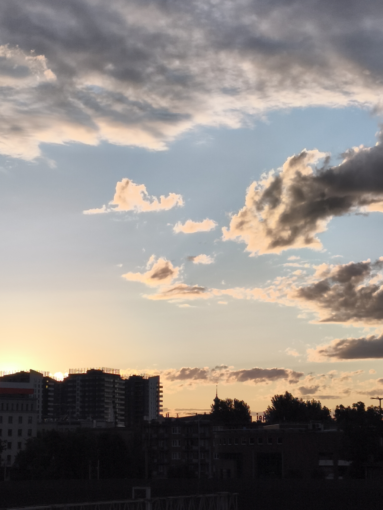 The image depicts a sunset over the city of Gdańsk, Poland. The sky is filled with dramatic clouds, illuminated by the orange and pink hues of the setting sun. In the foreground, the silhouettes of buildings and trees are visible against the sky. The buildings appear to be a mix of residential and commercial structures, with some under construction. The overall scene conveys a sense of urban tranquility as the day comes to a close. The lighting suggests it is either dawn or dusk.