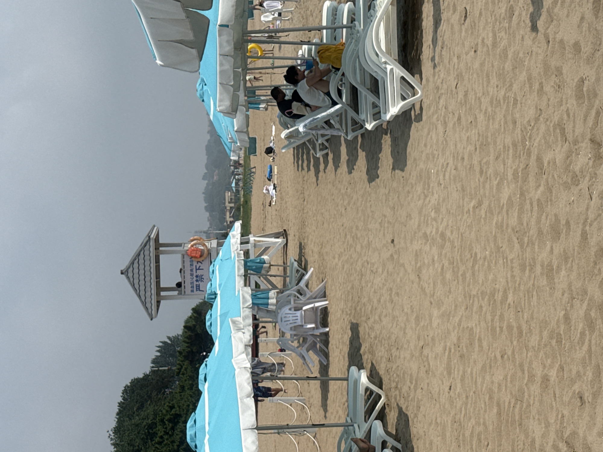 The image depicts a beach scene in Qinhuangdao, China, likely in the summer season due to the presence of beach umbrellas and loungers. In the foreground, rows of white plastic beach loungers are unoccupied, casting shadows on the sandy beach. Further back, a line of blue and white beach umbrellas provides shade. Several white plastic chairs are stacked near some of the umbrellas. A lifeguard tower, painted white with a grey roof, is visible in the middle ground. A person is seated inside the tower. A bright orange life preserver hangs on the side of the tower.
In the middle of the frame, under one of the umbrellas, a man and a woman are seated on loungers. The woman is looking down at something in her lap, while the man is looking to the right. A yellow bag is placed on the lounger next to them. Further in the background, other beachgoers are scattered across the sand, some lying on towels and others walking. Trees and some buildings can be seen on the distant horizon. The sky is overcast, suggesting a hazy or cloudy day. The overall impression is a moderately populated beach.
