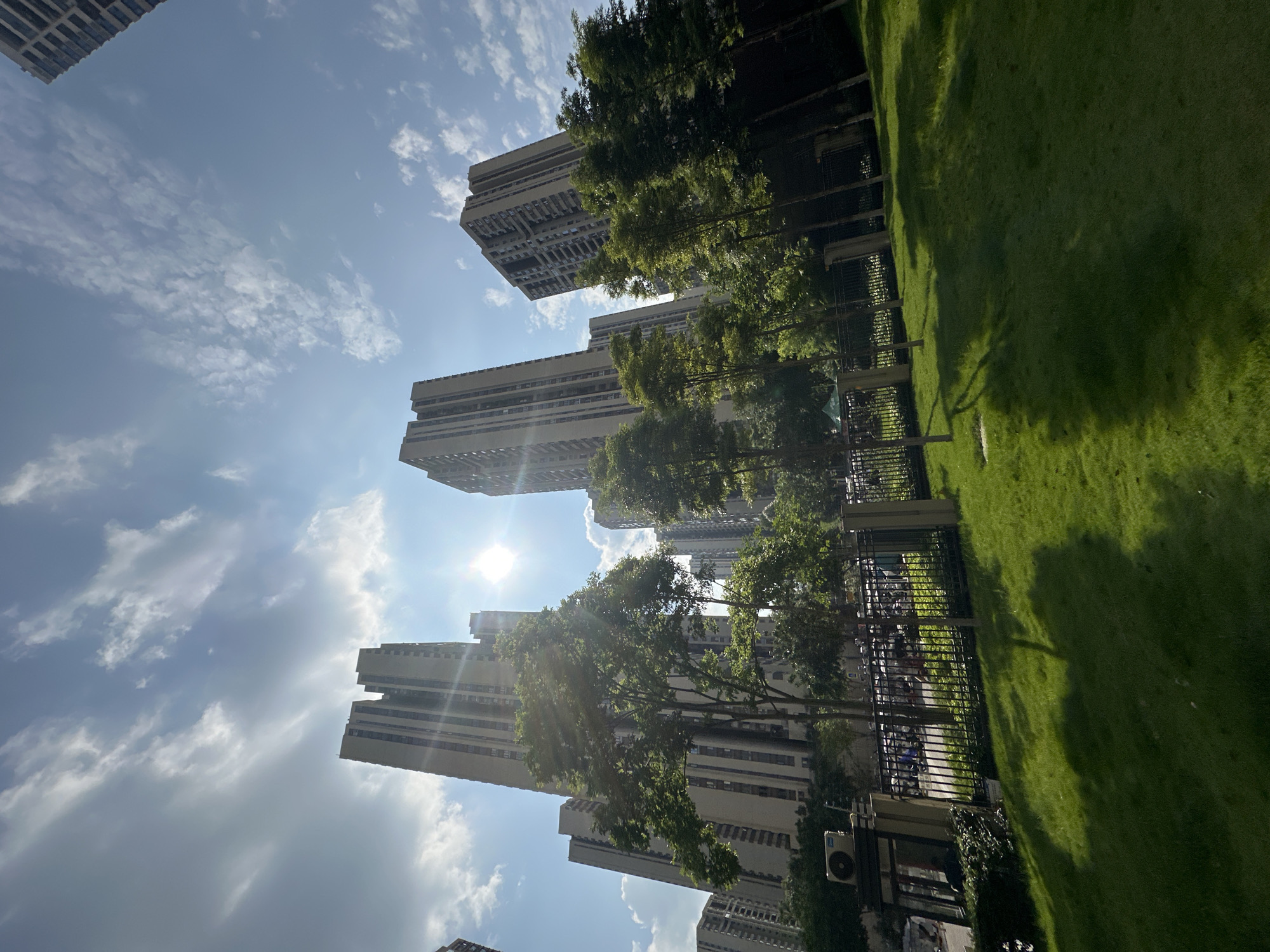 In a sunny daytime scene in Chongqing High-tech Zone, China, a cluster of tall apartment buildings rises against a bright blue sky with scattered white clouds. The sun, positioned prominently in the sky, casts a lens flare and illuminates the scene. In the foreground, a meticulously manicured green lawn slopes gently, with the dappled shadows of trees stretching across it. A black metal fence separates the lawn from a paved area behind it, where a row of trees stands, their leaves catching the sunlight. Beyond the trees and fence, several motorcycles and scooters are parked, suggesting a residential or communal area. The apartment buildings, characterized by their light-colored facades and numerous windows, are the dominant architectural elements, conveying a sense of urban density. An outdoor air conditioning unit is visible on a wall to the left of the frame.