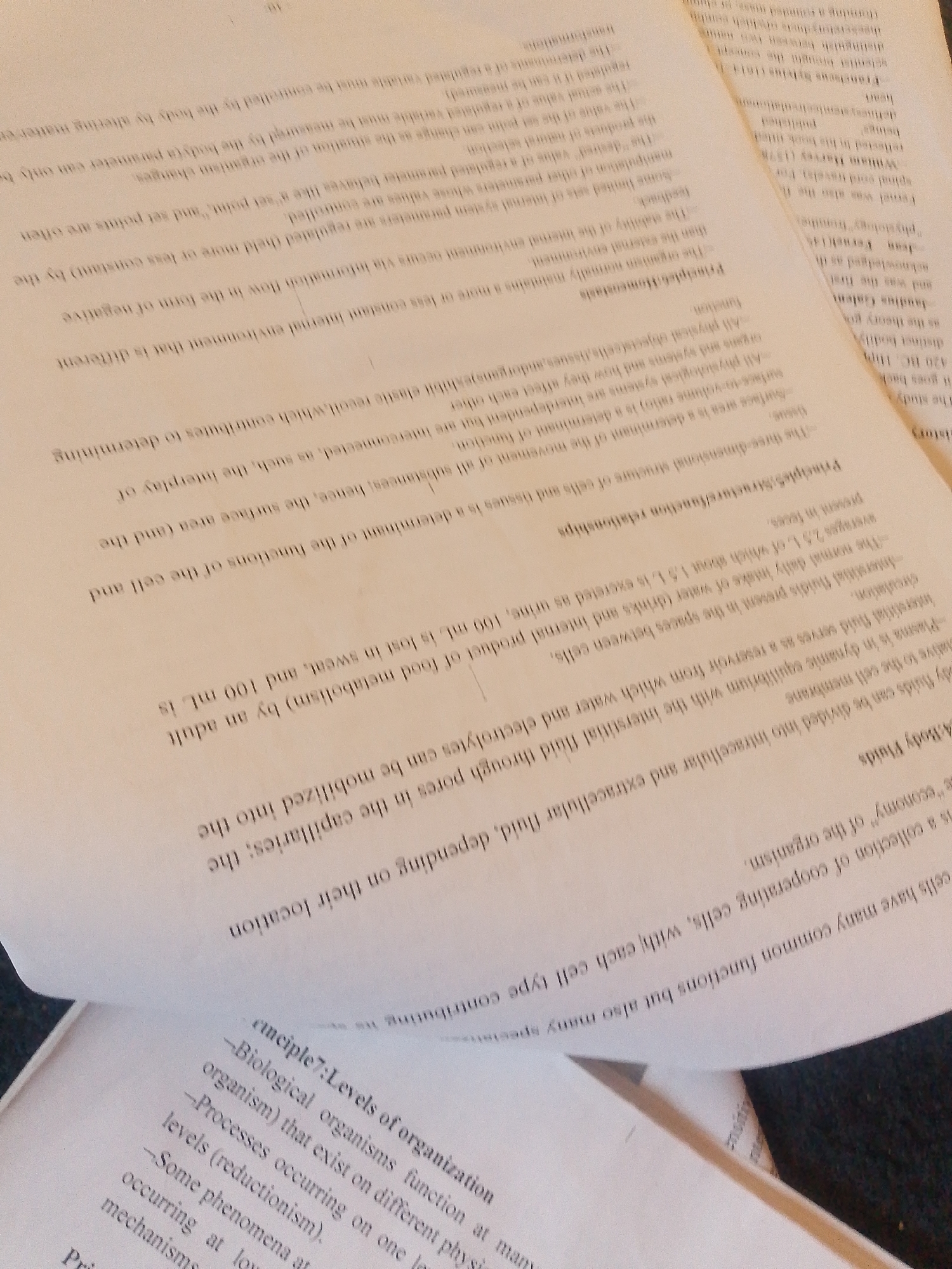 The image shows a close-up, overhead view of several pages of a textbook or study material, with one page significantly creased and obscuring some of the text. The pages appear to be aged, with a slightly yellowed hue. The text is printed in black ink and is in English.
The visible text discusses biological concepts such as "Body Fluids," "Principles: Structure/function relationships," "Principle6 Homeostasis," and "cmciple7: Levels of organization." Specific terms like "intracellular," "extracellular fluid," "plasma," "interstitial fluid," "negative feedback," "set point," and "biological organisms" are legible. There are also mentions of historical figures in science like "William Harvey" and "Galen."
The overall impression is one of academic study. The lighting suggests an indoor setting, possibly a desk or table where someone is reading or working. There are no people or specific objects other than the paper and ink. The creased nature of the pages and the angle of the photograph suggest that the material might be actively being used or reviewed. No specific time of day, weather, or emotional cues are discernible. The location context provided (Jalingo, Nigeria) does not offer any visual cues within the image itself.
