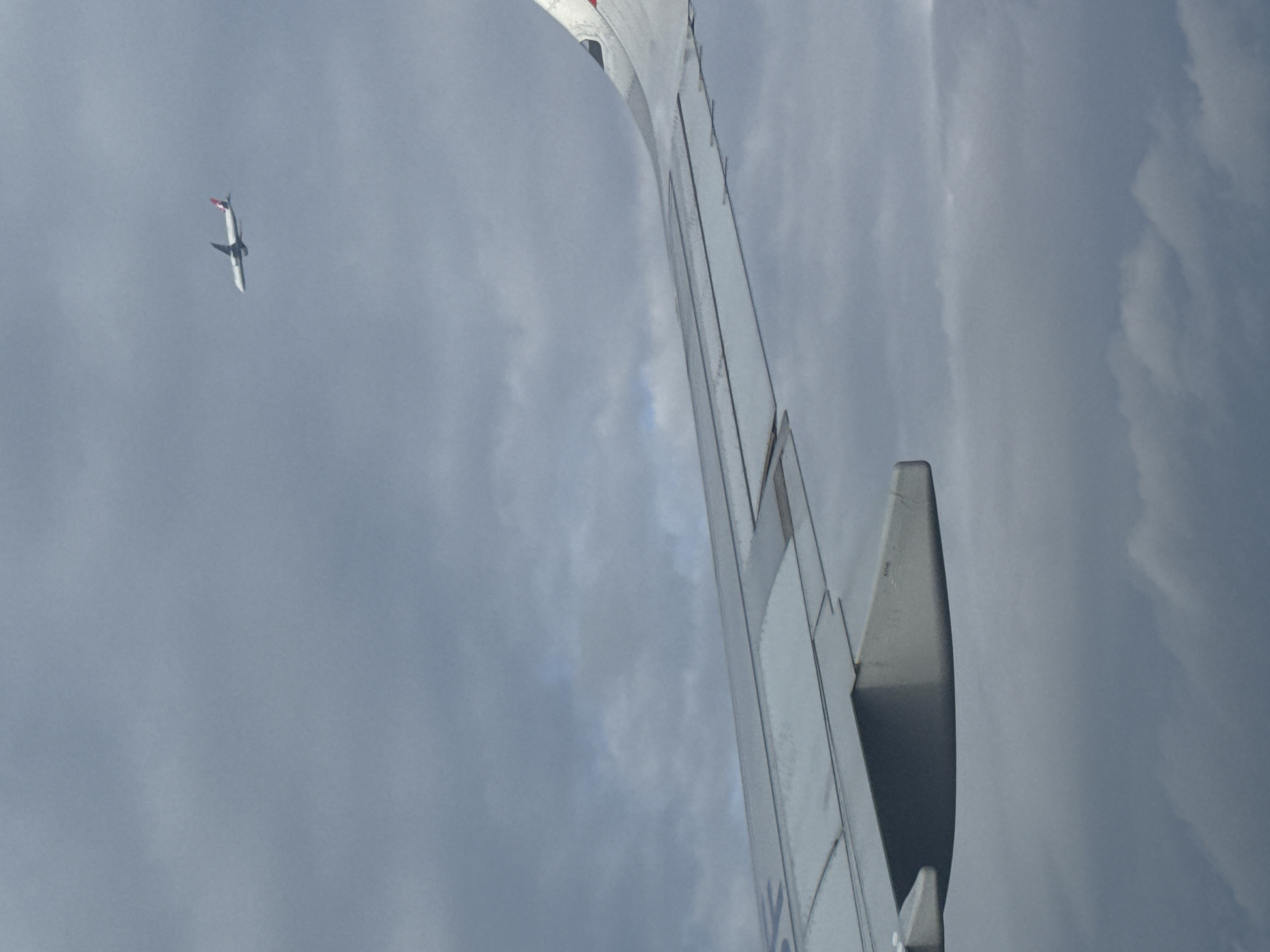 The image presents an aerial view, likely taken from an airplane window. A portion of the airplane's wing is visible in the foreground, with a grey, cloudy sky in the background. In the distance, another airplane is seen. Location cannot be determined from this image.