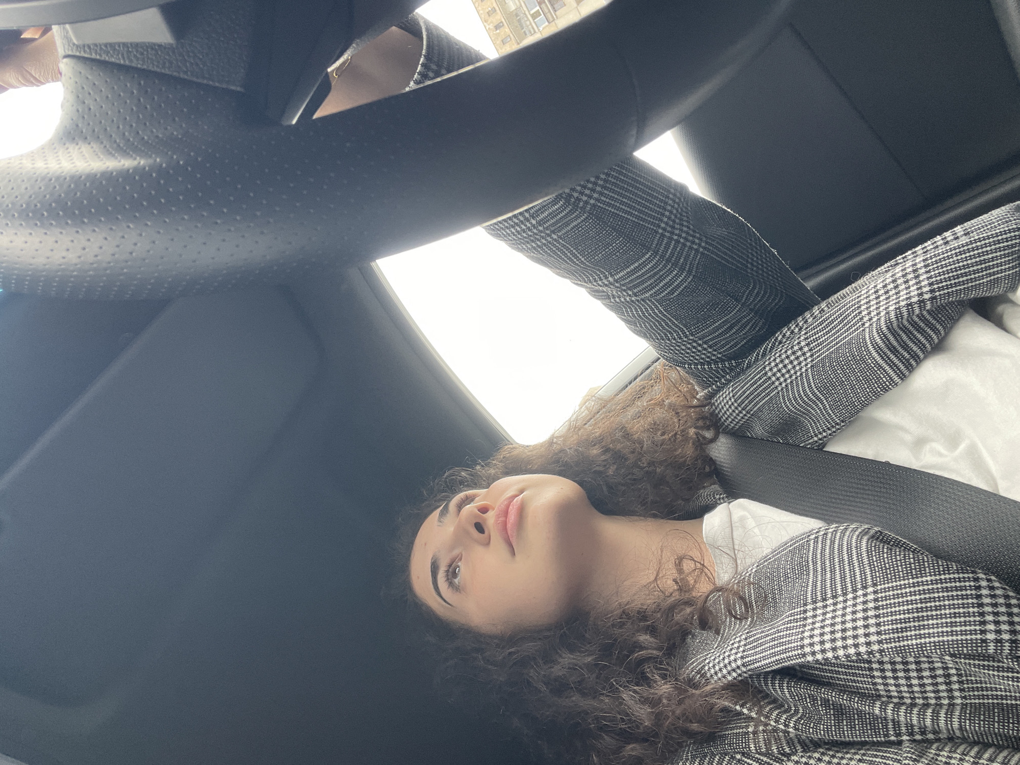 A woman with curly brown hair lies in the passenger seat of a car, wearing a black and white houndstooth blazer over a white shirt. She is secured by a black seatbelt, and her head is tilted upwards. The car's black dashboard and steering wheel are visible in the upper portion of the image. Through the windshield, an out-of-focus building can be seen, suggesting the scene is set within a city. Based on architectural styles and available information, the city is identified as Sofia, Bulgaria.