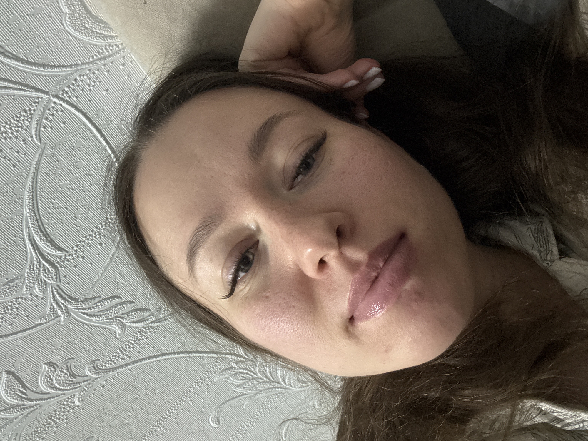 The close-up image, taken from a slightly tilted, overhead perspective, features a young woman reclining indoors. Her head and upper torso are visible, with her dark brown hair fanned out around her. She looks directly into the camera with a neutral, slightly contemplative expression, her lips parted and glossy with a pinkish tint. Her dark eyes are enhanced by prominent eyelashes, and her right eye (viewer's left) appears subtly narrowed. Her facial skin displays natural texture, including visible pores and some scattered freckles. One hand is visible near her head, showcasing short, white-painted fingernails.
The background on the left side features a light-colored wall or wallpaper with a raised, patterned texture, possibly a floral or vine motif. On the right, behind her head, a soft, light-colored cushion or pillow is visible. The lighting is soft and even, typical of an indoor environment. There is no visible text within the image. The provided location context is Ферма, Russia.