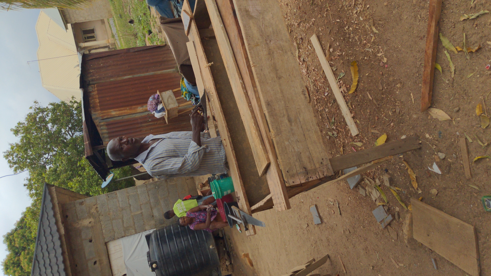 The image, rotated 90 degrees clockwise, depicts an outdoor scene featuring an adult man, an adult woman, and a child engaged in daily activities. In the foreground and mid-ground, an adult man, wearing a striped shirt, is focused on working with large wooden planks laid out horizontally. Wood shavings are visible on and around the planks, suggesting he is engaged in woodworking, possibly planing or shaping the timber.
To the man's left, an adult woman in a patterned dress is seated on a low object, possibly a stool, holding a small item in her hands. Beside her, a child, wearing a yellow t-shirt with the word "BOY" faintly visible on it, is also seated. Nearby, a large black water tank, green and red buckets, and a small satellite dish are present.
The setting combines elements of construction and residential living. Behind the man, there is a structure made of corrugated metal sheets. Further into the background, a partially built concrete block wall is visible, along with a more complete residential building featuring a light-colored, multi-gabled roof. Lush green trees flank the left side of the image, and the ground is dusty earth, scattered with dried leaves, small pieces of wood, and debris. The scene is illuminated by natural daylight, indicating it is daytime. The environment suggests a semi-urban or rural area, consistent with the context of Masaka, Nigeria, showing an informal workspace alongside living quarters.