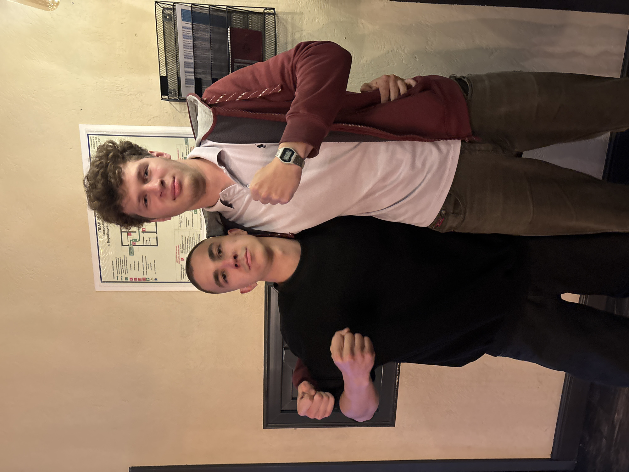 Two men are posing for a photo in what appears to be an indoor establishment in Birobidzhan, Russia. The man on the left is shorter, wearing a black t-shirt and black pants. The man on the right is taller, with curly hair, wearing a light-colored polo shirt and a maroon zip-up hoodie. Both men have their fists clenched. Behind them, on the wall, is a document, possibly a fire safety plan, with Russian text. To the right of the document, a black metal rack holds several papers. The wall behind them is a light tan color, and to the left, there is a dark framed rectangular object, possibly a fireplace or a decorative element.