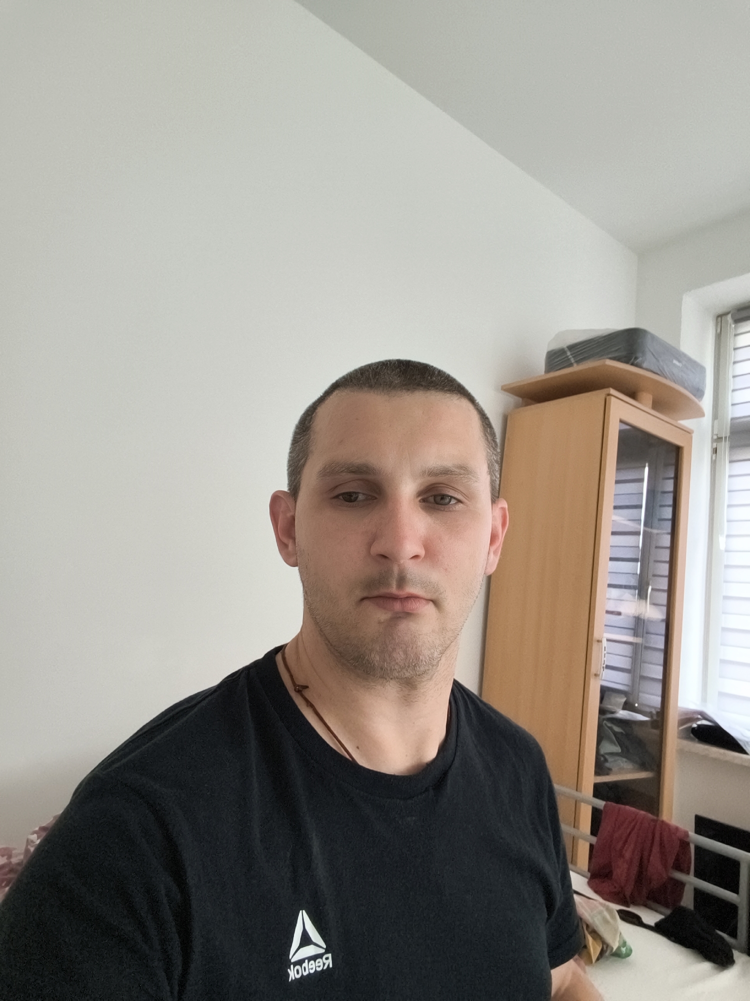 A man with short, dark hair and a beard is pictured in a selfie. He is wearing a black t-shirt with a white Reebok logo. He is standing in front of a plain white wall. To his right, a wooden cabinet with glass doors is visible, and a black printer is on top of it. A window with blinds is to the far right. There is a metal bed frame with some bedding and clothing in the background. The overall environment appears to be an indoor space, possibly a bedroom or living area, located in Wolgast, Germany.