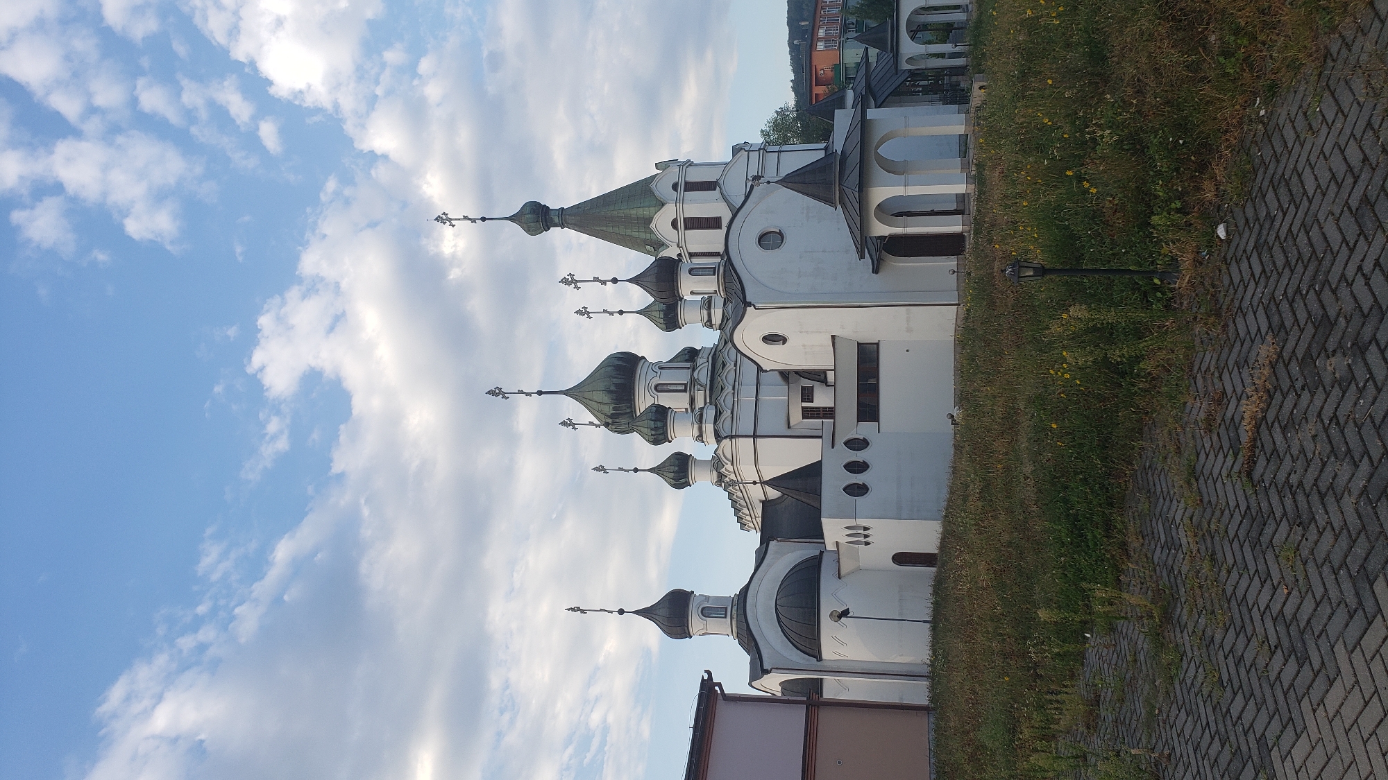 The image shows an exterior view of the Holy Trinity Orthodox Church in Prešov, Slovakia. The church is a white, multi-domed structure with green spires topped with crosses. It is situated on a slightly overgrown grassy area, with a paved walkway in the foreground. The sky above is partly cloudy, with blue sky visible. A street lamp is visible in the grass to the right of the walkway. In the background, a building with an orange roof is visible on a hillside. There are no people or animals present in the image. The scene appears to be captured during daylight hours.