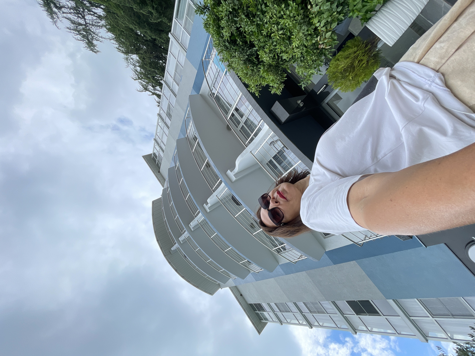 An extreme low-angle, wide-shot selfie shows a woman with dark hair and sunglasses, wearing a white t-shirt and light-colored pants, standing in front of a tall, modern, light blue and grey building in Abaata, Abkhazia. The building has multiple curved balconies with silver railings. To the right of the woman is a vibrant green shrub in a white planter, and further back, a taller evergreen tree. The sky above is overcast with patches of blue peeking through. The perspective emphasizes the imposing height of the building and creates a dramatic, slightly disorienting feel.