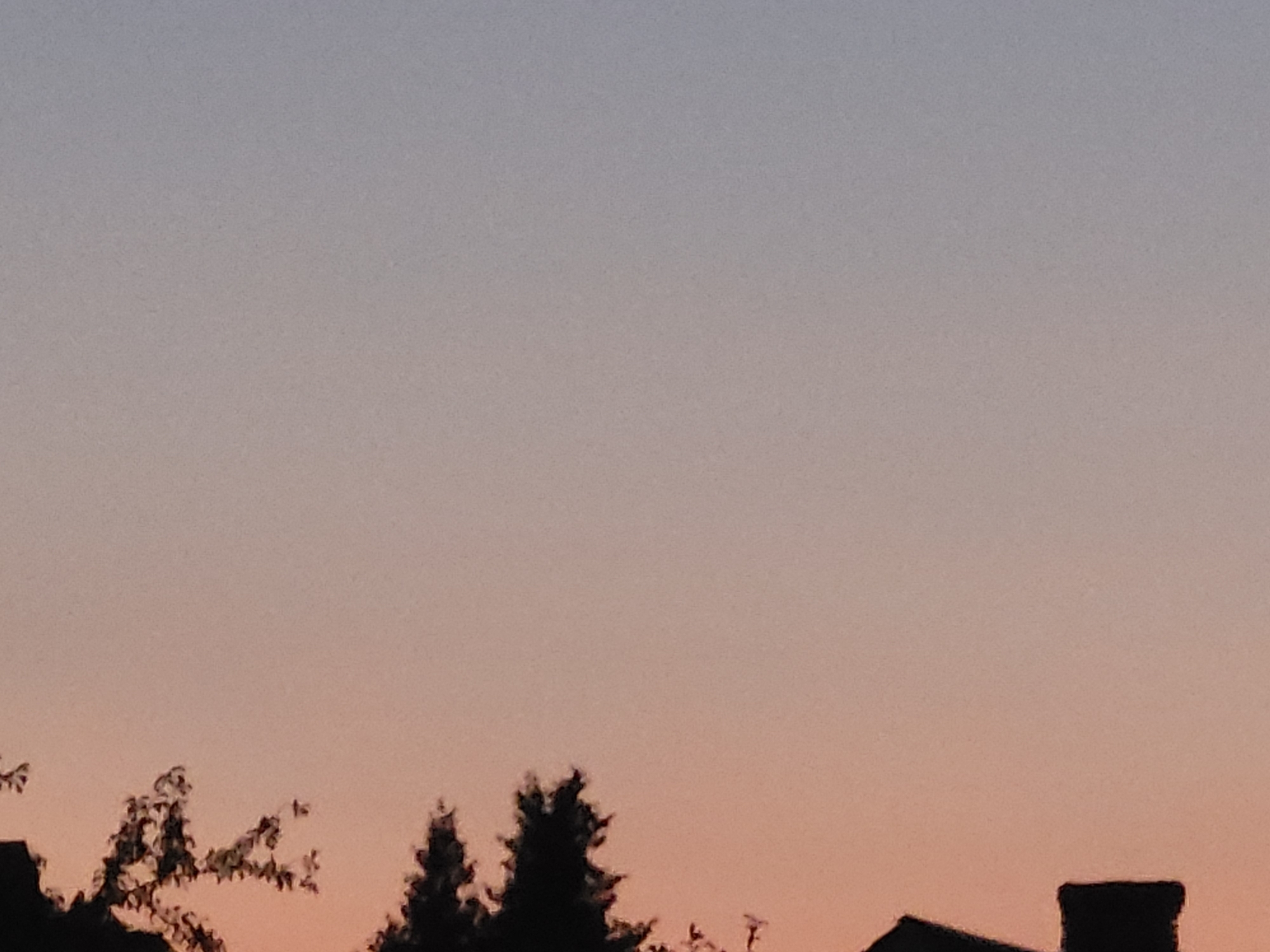 The image captures a silhouetted view of treetops and a portion of a building's roof with a chimney against a gradient sky that transitions from a light blue at the top to a warm peach/orange hue at the bottom, indicative of either sunrise or sunset. The location is identified as Ozolnieki, Latvia. There are no people or discernible actions depicted in the image. The focus is on the natural light and the dark silhouettes of vegetation and architecture against it.