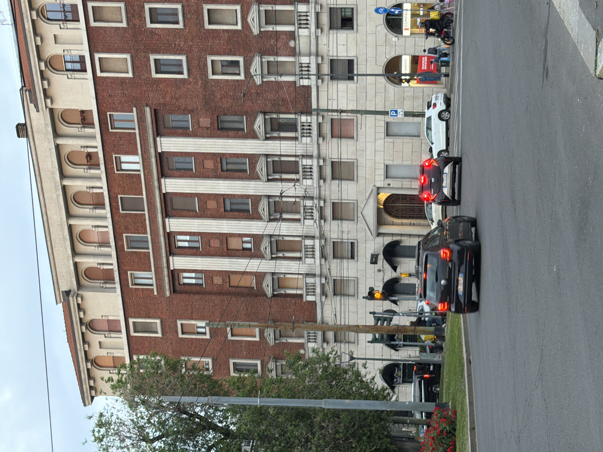 The image depicts a street scene in Milan, Italy. A multi-story building with a brick and stone facade lines the left side of the frame. The architecture suggests a classic Italian style. The street is moderately busy with vehicular traffic, including cars and scooters. Pedestrians are visible in the background, near a sign that has the name "Compass". Streetlights and traffic signals are also present. The scene captures a typical urban environment in Milan.
