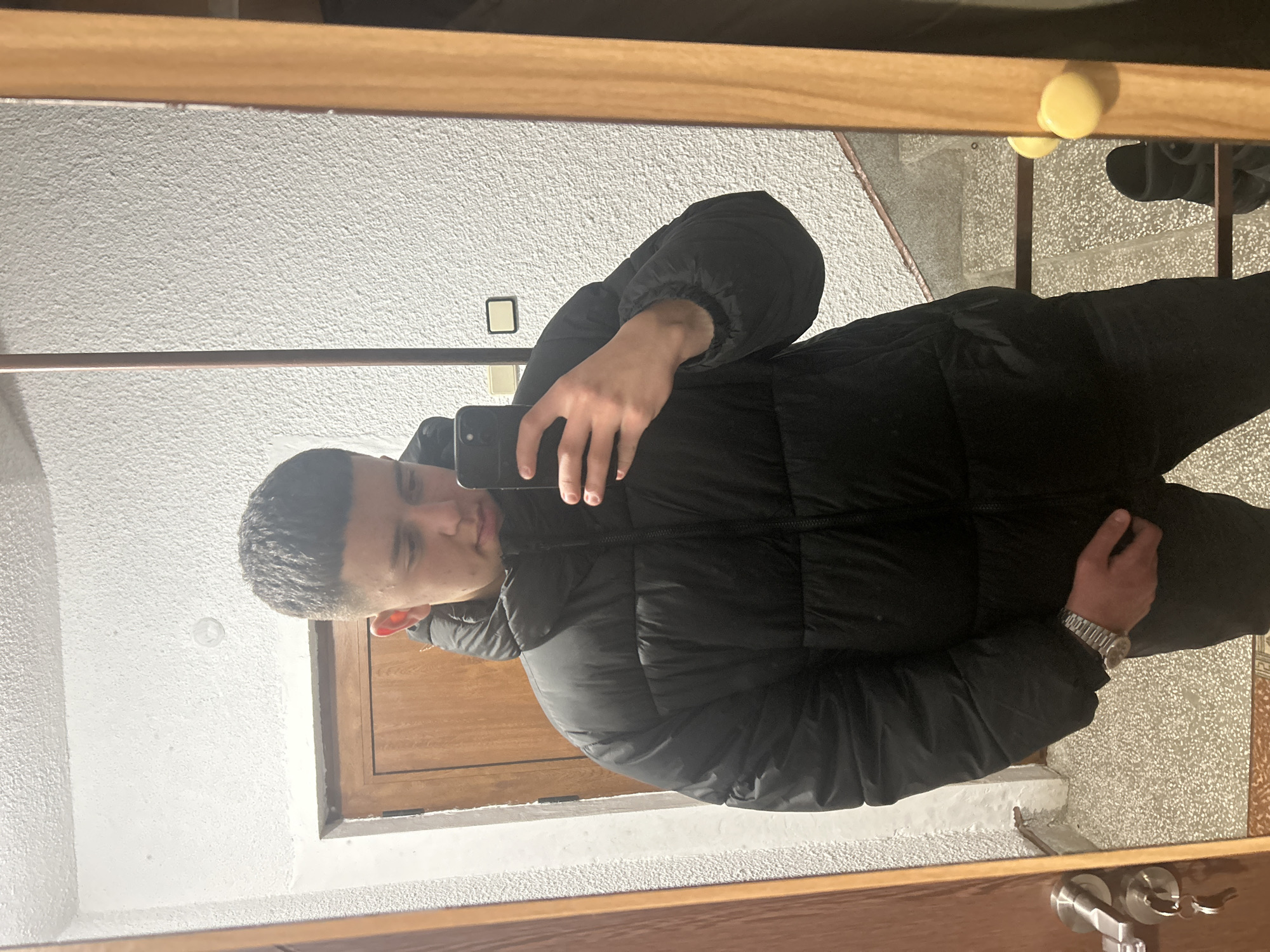 A young man is taking a selfie in a mirror. He is wearing a black puffer jacket and dark pants. A wristwatch is visible on his left wrist. The background shows a white textured wall, a wooden doorframe, and a glimpse of a tile floor. Based on the query, the location of the image is assumed to be in the city of Zlatograd, Bulgaria.