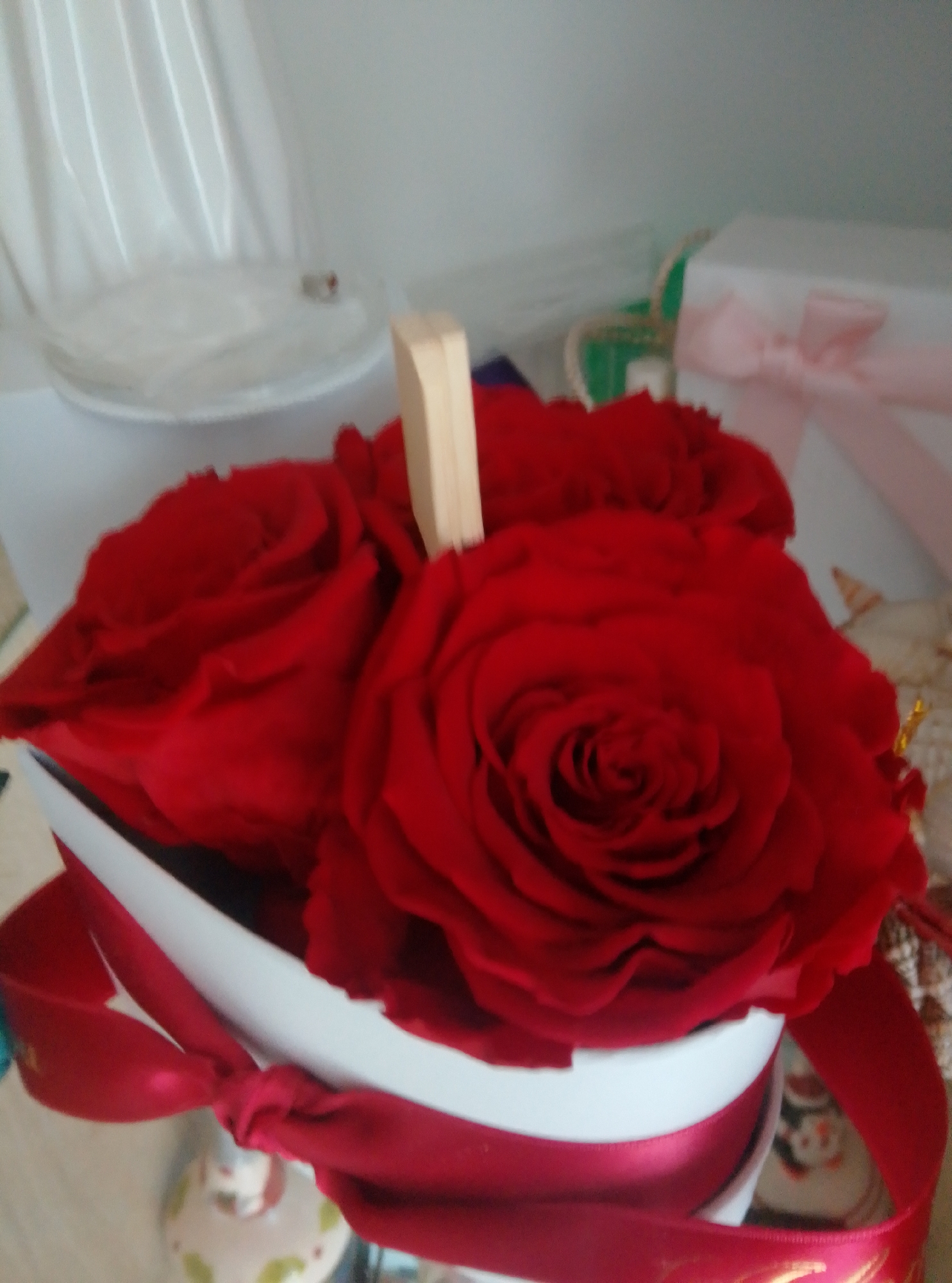 A close-up, high-angle shot depicts a bouquet of red roses in a white box, adorned with a red ribbon. A wooden stick protrudes from the arrangement. In the background, a white gift box with a pink bow and a translucent white object are visible. The scene is brightly lit, with the overall impression being one of a gift or decorative item. The image quality is slightly blurred, suggesting it was taken in a home or indoor setting, not outdoors. There is no discernible information to place this scene in Wedel, Germany or any specific urban location.