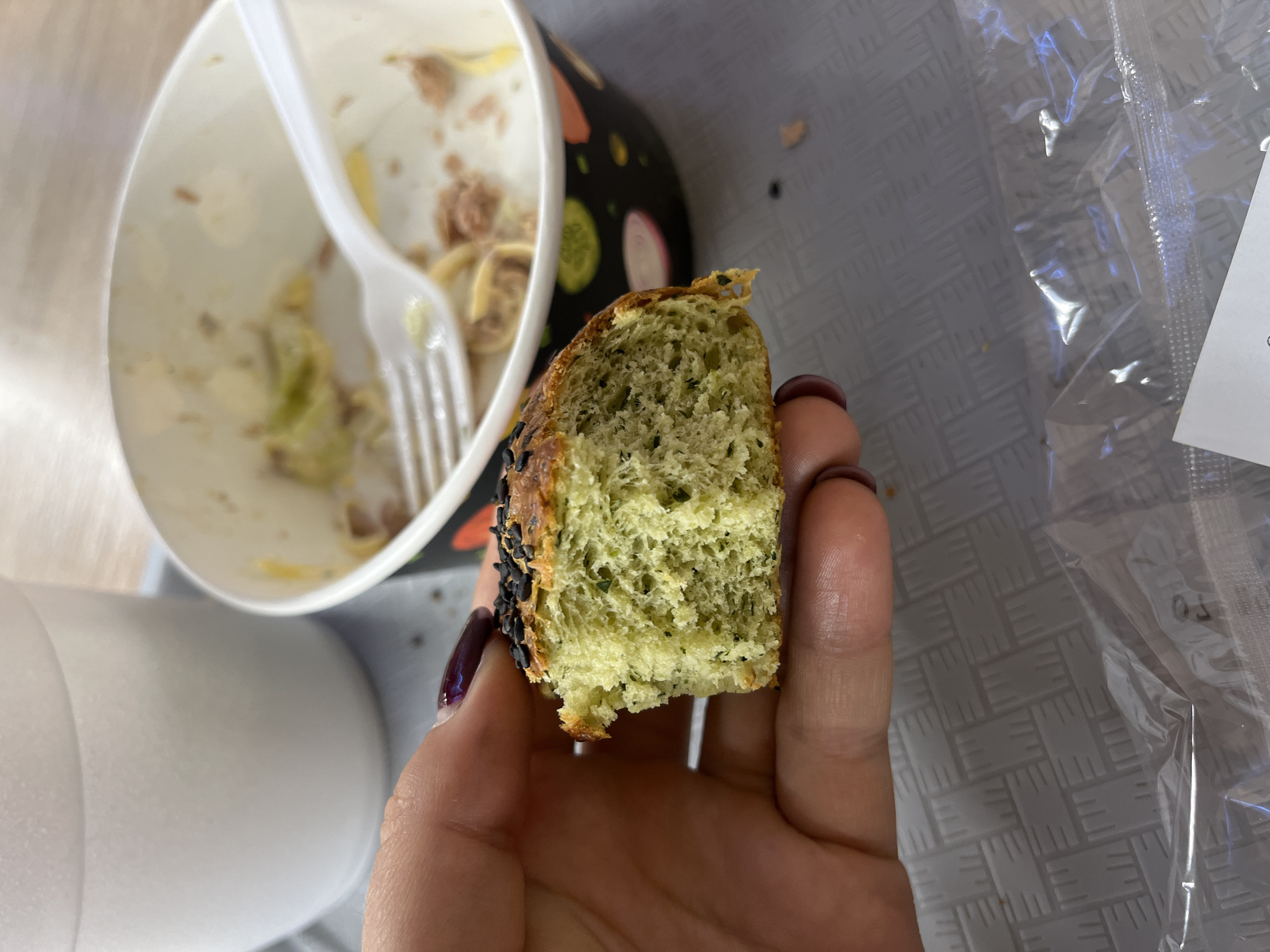 In this close-up shot, a person's hand, with dark purple nail polish, holds a piece of green bread topped with black seeds. The bread appears to be a savory type, possibly containing herbs, and has a slightly crumbly texture. In the background, a white plastic cup and a partially eaten bowl of food with a plastic fork are visible. The setting is likely a cafeteria or dining area, as suggested by the plastic tray and the disposable tableware. The scene offers no direct indicators to pinpoint its location within Minsk, Belarus.