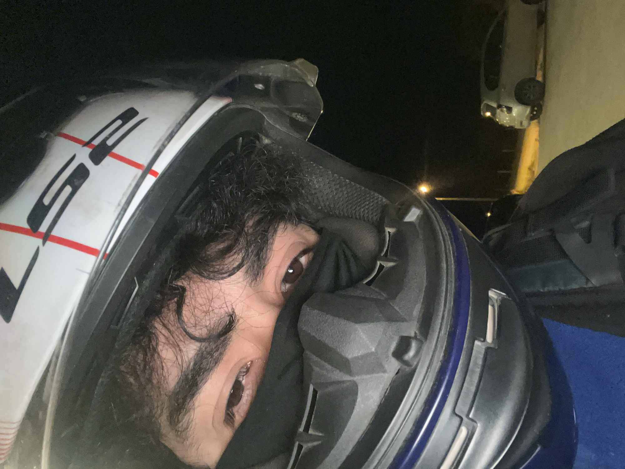 A person wearing a motorcycle helmet is photographed close-up at night. The helmet is primarily white with black and red accents. The person's eyes are visible; they are partially obscured by a black garment covering part of the face. The background is dark, but a vehicle is visible in the upper portion of the frame. There are no indicators to confirm the image's location in Ushuaia, Argentina.