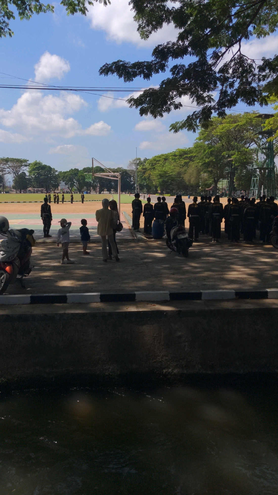 In the city of Magelang, Indonesia, this image captures a group of people in an outdoor space, likely a field or court area with a basketball hoop visible in the background. In the foreground, two children are standing near two adults. To the right, a larger group of individuals, many in matching dark uniforms, are gathered. Several motorcycles are parked in the area. The environment is sunny with scattered clouds, and the scene is bordered by trees and a dark, water-filled channel in the very front.