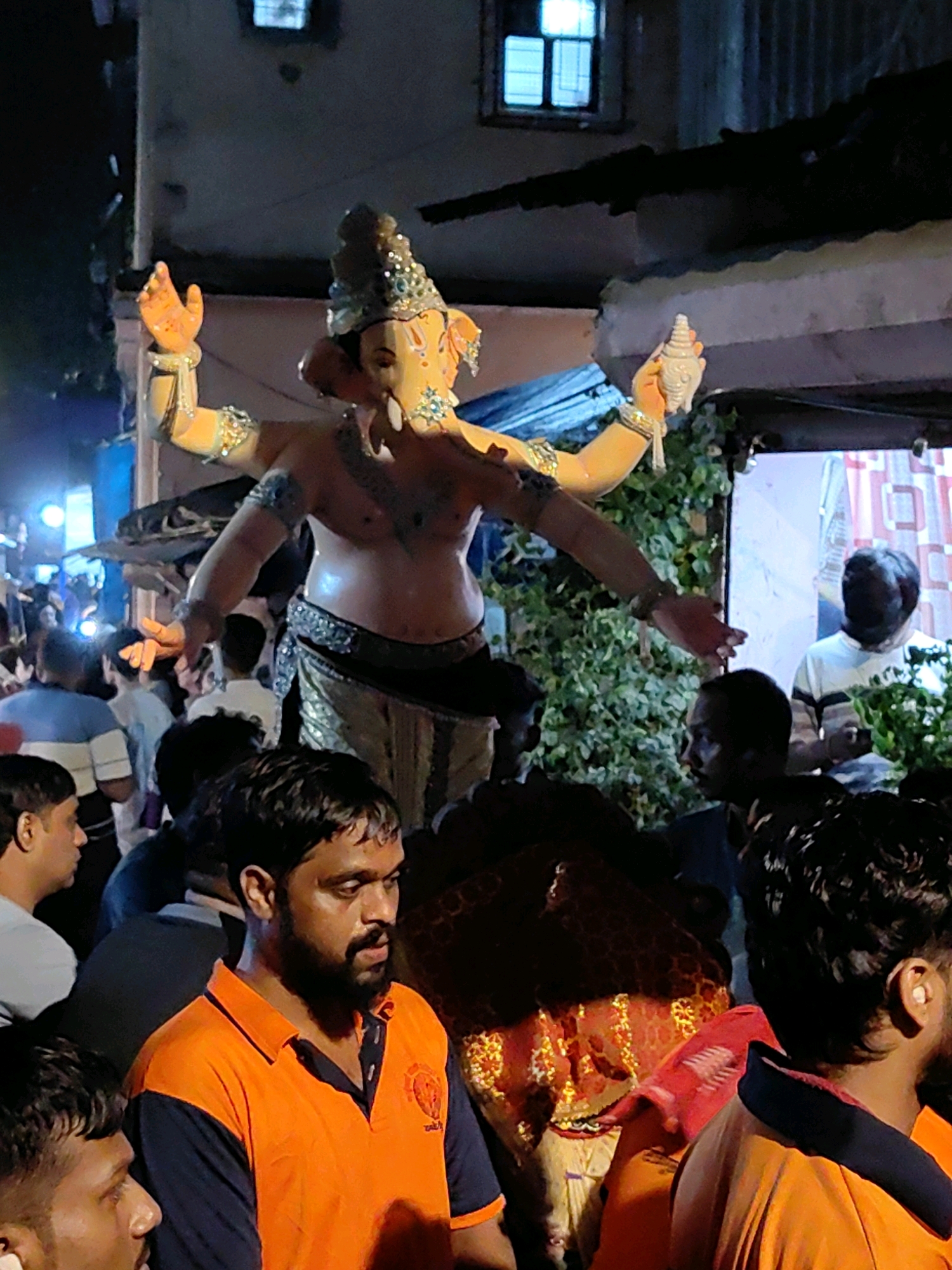 This is a medium shot, eye-level, vertical photograph taken at night in Mumbai, India, during a religious procession. The central subject is a large, ornate statue of the Hindu deity Ganesha, depicted with multiple arms and adorned with jewelry and a crown. The statue is carried on a platform, surrounded by a crowd of people. In the foreground, a man with a beard wearing an orange and navy blue polo shirt is looking to the right, his gaze directed away from the camera. Behind him, the shoulders and heads of other individuals are visible. The background shows a building with a window and foliage, illuminated by artificial lights, suggesting an urban setting. The overall atmosphere is festive, indicative of a cultural or religious event.