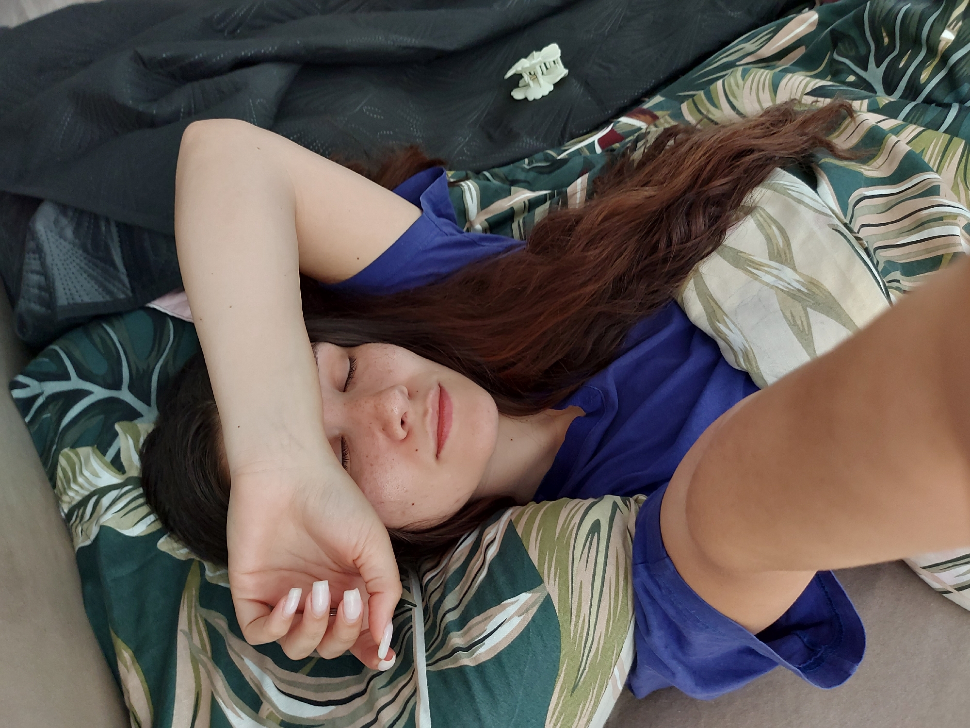 A woman is depicted lying in bed, appearing to be asleep or resting. Her eyes are closed, and her left arm is draped over her forehead and face, partially shielding her eyes. She has long, wavy dark brown hair spread out on the pillow and duvet. She is wearing a simple blue t-shirt. Her face shows light skin with visible freckles on her nose and cheeks, and her lips have a natural pink hue. Her fingernails are manicured with a light, opaque polish.
The bed is covered with two distinct types of bedding. On the left and behind her head is a dark grey, textured or quilted blanket or duvet. To her right and beneath her head is a duvet or pillow with a vibrant tropical leaf pattern in shades of green, white, and beige. A small, white, claw-style hair clip rests on the dark grey bedding near the top left of the frame. The lighting in the image suggests it is daytime. The overall scene conveys a sense of peaceful slumber or quiet relaxation in a bedroom setting, located in Shumen, Bulgaria. No visible text is present in the image.