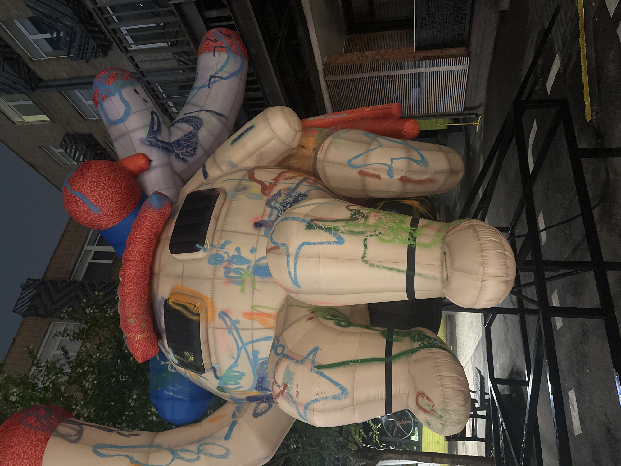 An inflated, multi-colored sculpture stands on the street in Moscow, Russia. The sculpture has a white base with blue, green, orange, and red graffiti markings. It also features red and blue spherical and cylindrical shapes, and black accents that appear to be windows. The sculpture is situated near a building with visible windows and architectural details. A metal scaffolding or frame is visible near the sculpture, and the surrounding area includes a street with painted lines. The time is dusk or evening based on the ambient lighting.