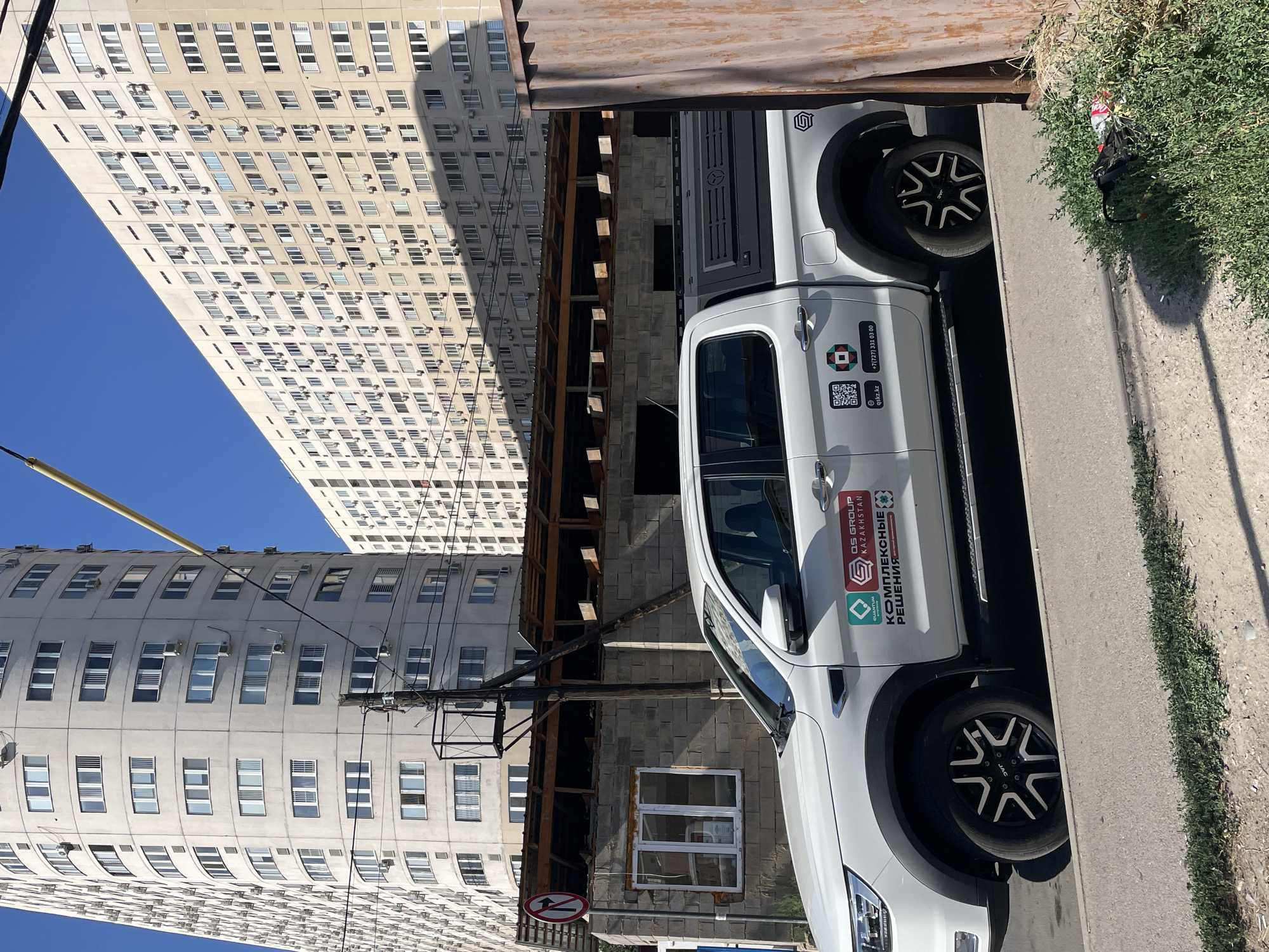 A white pickup truck with a black canopy is parked on a street in Almaty, Kazakhstan, in front of a construction site and two tall apartment buildings. The truck has several stickers on its side, including one with the text "QS GROUP KAZAKHSTAN" and another with "КОМПЛЕКСНЫЕ РЕШЕНИЯ". A traffic sign indicating a right turn restriction is visible on the left side of the image. The apartment buildings are modern, with many windows and air conditioning units. The sky is clear and blue. There is some construction debris and a rusty metal fence on the right side of the image.