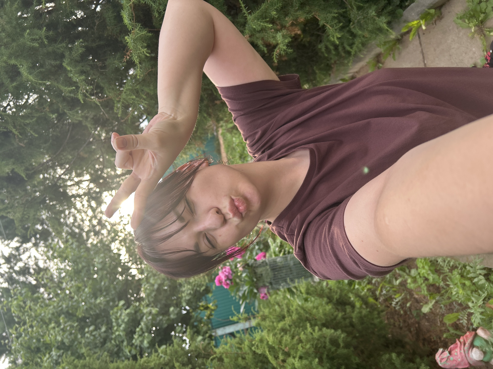 A young woman with dark hair, wearing a maroon t-shirt, is taking a selfie outdoors in a garden setting. She is puckering her lips and holding up two fingers to her forehead in a peace sign. The background shows lush green foliage, including several coniferous trees and some pink flowers. A glimpse of a green fence and a small garden gnome can be seen on the left side of the image. The sun appears to be low in the sky, casting a warm light and creating some lens flare. The location is identified as Фоки, Russia.