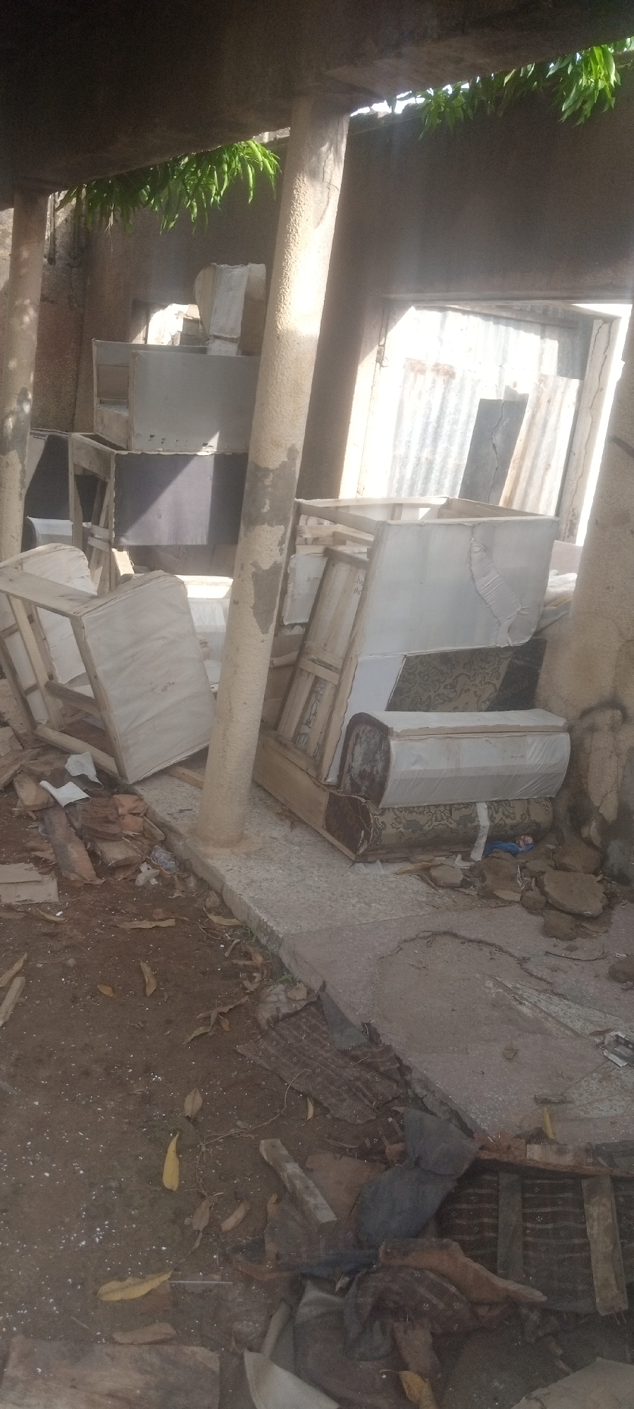 The image depicts a cluttered, possibly abandoned outdoor space in Sabon Tasha, Nigeria. There are several pieces of discarded furniture, including upholstered chairs and what appear to be parts of sofas, piled up near weathered concrete pillars. The ground is littered with debris, including wood scraps, leaves, and other unidentifiable materials. The background shows a corrugated metal structure or wall. The overall impression is one of neglect and disuse.