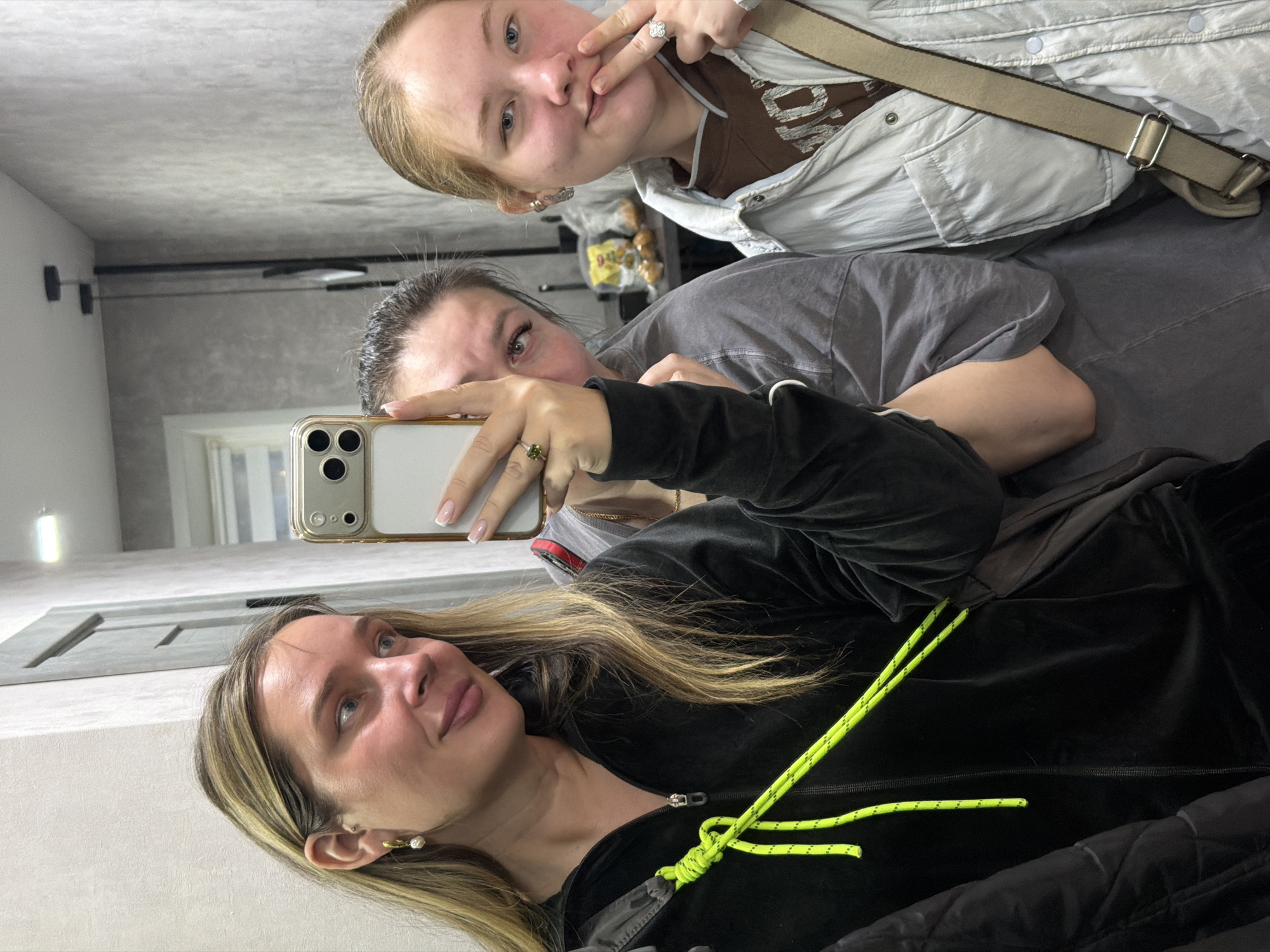 Three females are taking a selfie in what appears to be an interior space with neutral-toned walls. The foreground shows a woman with blonde highlights, wearing a black zip-up jacket with neon green drawstrings. Her face is angled slightly upwards, and she's looking to the side. Behind her, another woman holds up a phone to capture the image. A third, younger female is in the background, pulling a face and making a hand gesture. The clothing worn by the background women seems casual, with a gray top and a light-colored jacket visible. The location of the city of Chaikovsky, Russia, cannot be determined from the visual information provided in the image.