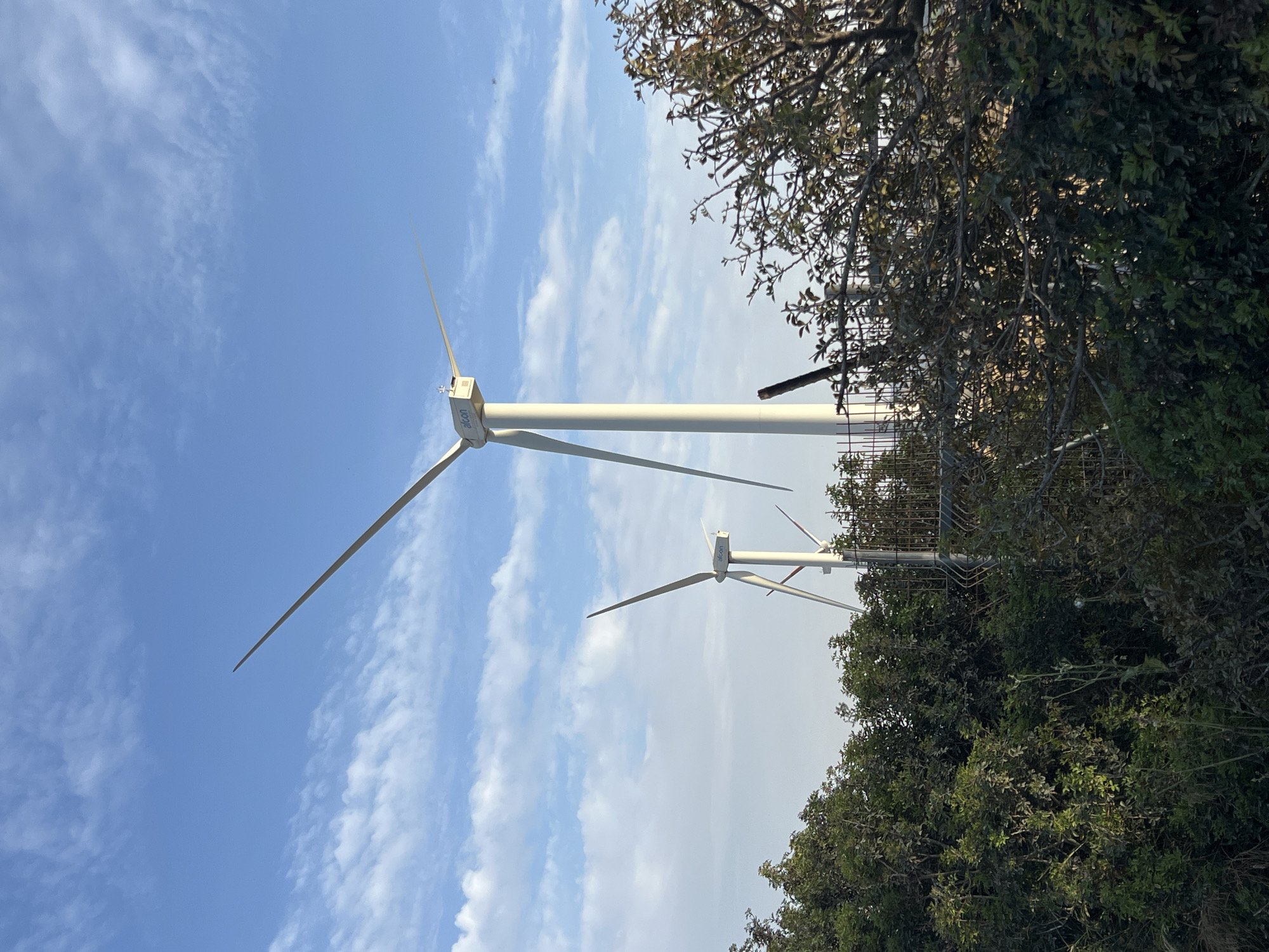 The image shows two wind turbines in Emek HaMaayanot Regional Council, Israel. The sky is blue with scattered white clouds. A tree and foliage partially obscures the view of the turbines and a metal fence is visible near the trees. The turbines are white, with three blades each. The logo "Aicon" is visible on one of the turbines.