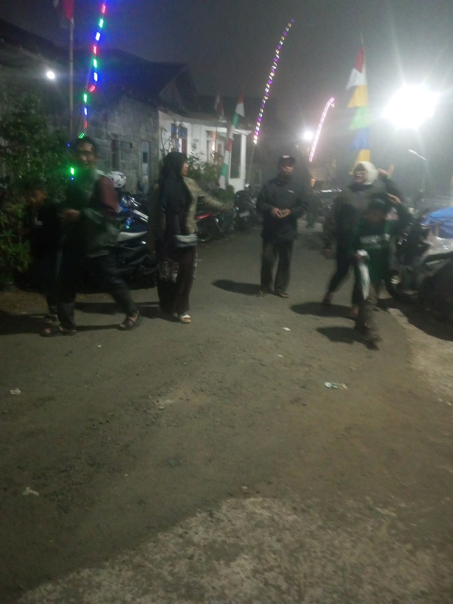 The image depicts an outdoor scene at night, likely in a residential area of Keseneng, Indonesia, given the context. The setting is a paved road with some sparse vegetation on the left. Several individuals are present, appearing to be engaged in a casual gathering.
In the foreground, a man in a dark jacket and a baseball cap stands facing the camera, his hands clasped. To his right, a woman in a dark headscarf and a patterned skirt is visible. Further to the left, a person in a maroon shirt and dark pants is partially obscured by the frame. Behind them, several motorcycles are parked, suggesting a common mode of transportation.
The background features buildings with illuminated windows, indicative of a residential or commercial structure. Decorative lights, including a string of colored lights (red, green, blue), are strung along a pole, and another string of purple lights curves upwards, suggesting a festive occasion or celebration. A brightly lit light source on the right side of the frame creates a lens flare effect, partially obscuring details in that area. The overall atmosphere is dimly lit, with a slight haze or fog contributing to the nighttime ambiance. The ground appears to be asphalt or a similar paved surface.