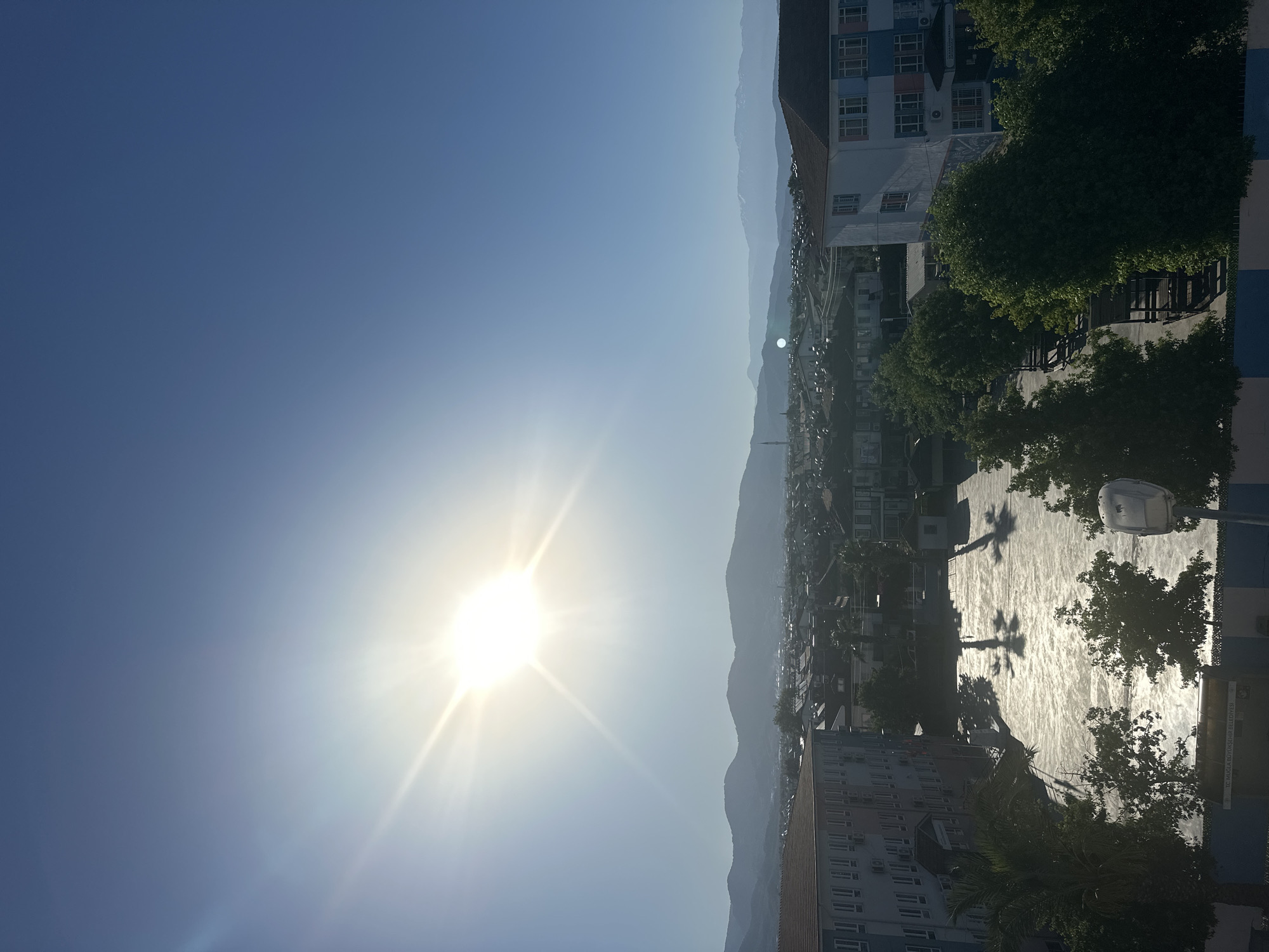The image shows a view of Fethiye, Turkey, looking towards a mountain range in the distance. The sun is bright in the sky, with lens flare visible. A cityscape is visible to the right. In the foreground is a building. There is a paved area reflecting sunlight. Trees and street lights surround it. Shadows of the trees are also visible on the pavement. Buildings of various sizes are seen along the street.
