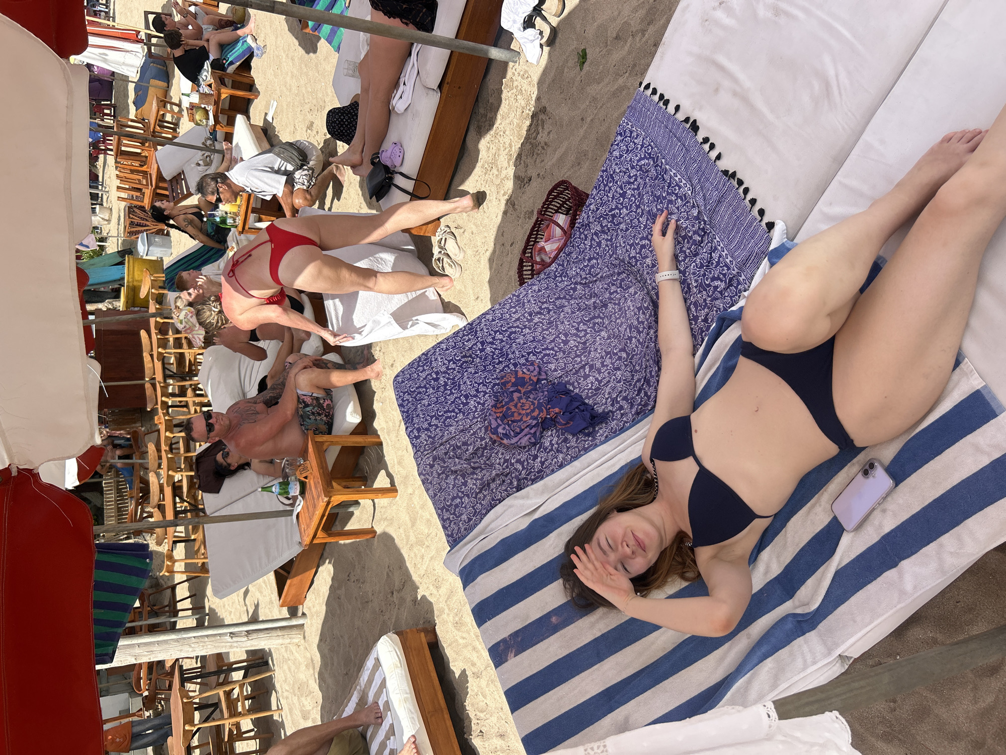 In Kuta, Indonesia, a group of people are relaxing on a beach under umbrellas and tents. In the foreground, a woman in a black bikini lies on a beach chair with a blue and white striped towel, shielding her eyes from the sun. A light purple phone rests on the towel next to her. To her right, another beach chair is covered with a purple and white patterned throw. Behind her, a group of people are seated at wooden tables and chairs, some with drinks. In the midground, a man and woman in swimwear are conversing. Further back, more people are seated on lounge chairs. The ground is covered in sand.