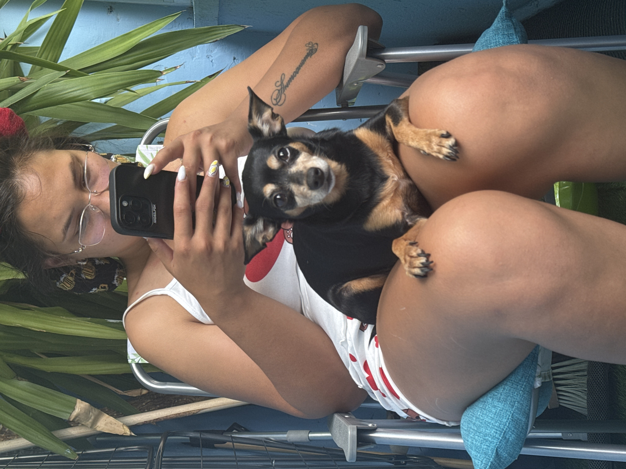 A woman is sitting outdoors on a chair, holding a smartphone and looking at it. A small dog is sitting on her lap, facing forward. The woman is wearing a white tank top with red heart patterns and white shorts with red trim. She has on glasses and her hair is pulled back. The dog is black and tan with floppy ears. In the background, there are green plants and a blue wall. A tattoo is visible on the woman's arm. The scene appears to be a casual, outdoor setting.