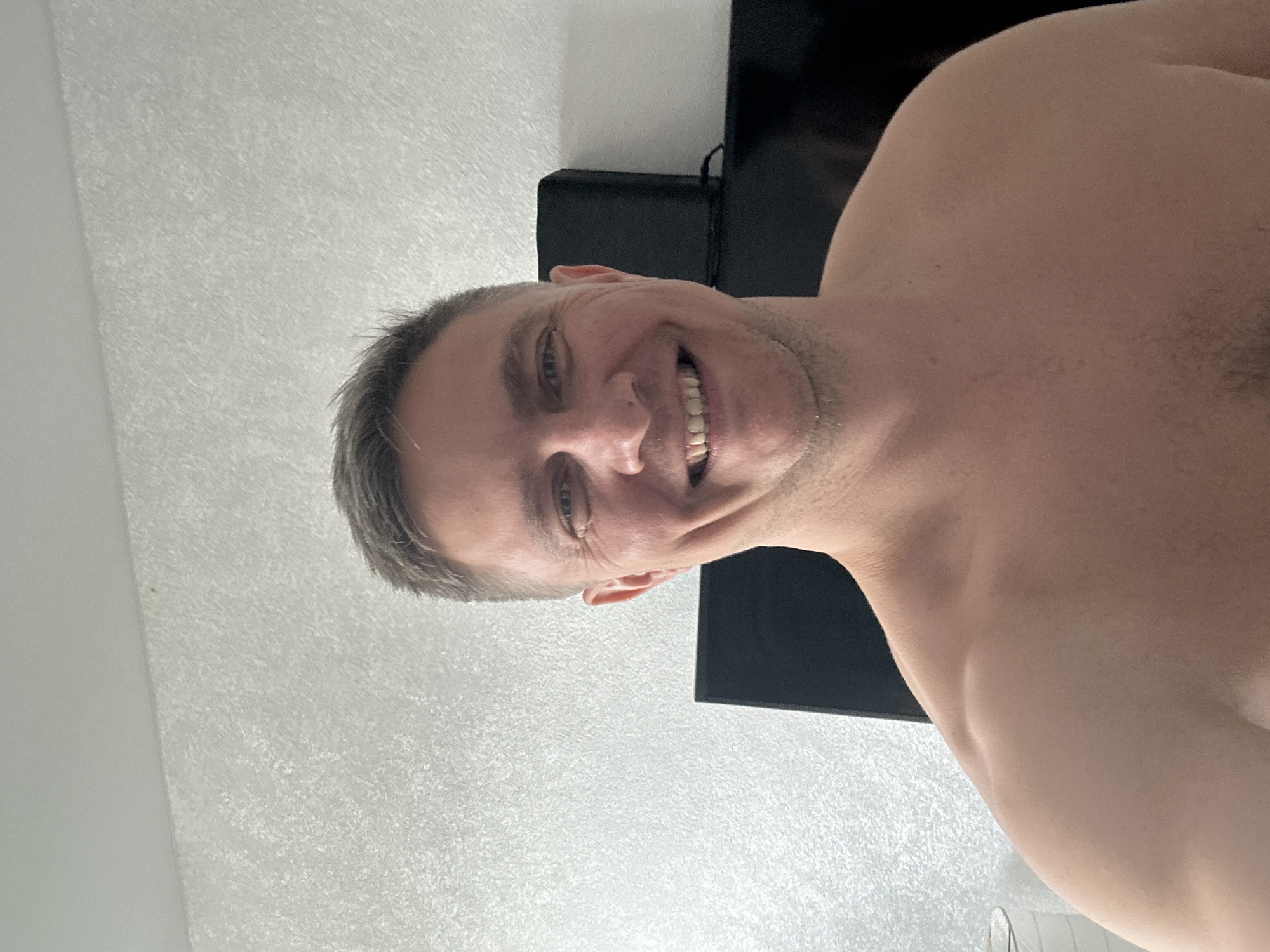 A shirtless man is smiling at the camera. He has short grey hair, fair skin, and some stubble. A black TV is partially visible behind his head. The background appears to be a white textured wall. The image was taken indoors. There is no information available to conclude that this image was taken in Бараўлянскі сельскі Савет, Belarus.