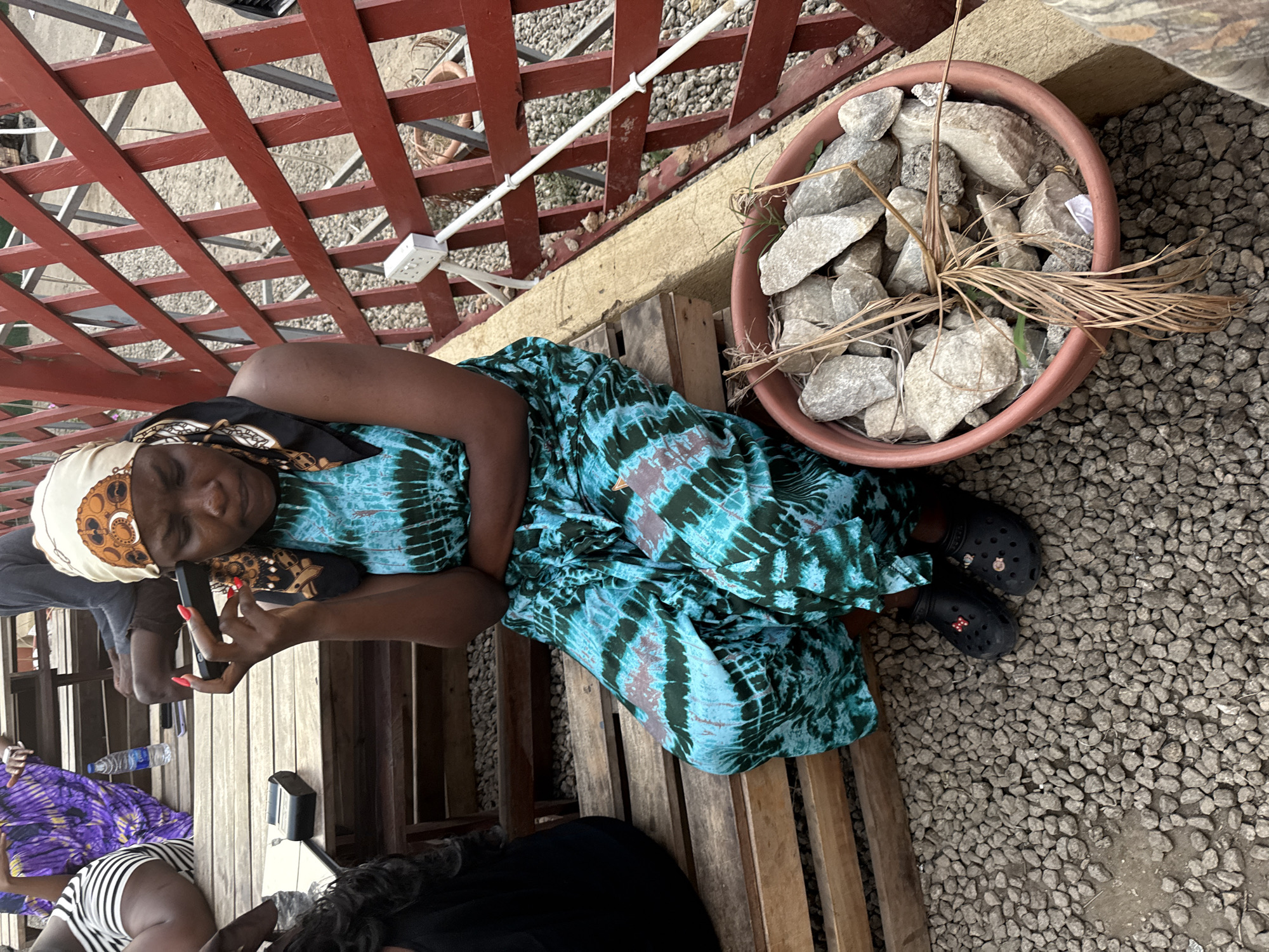 A woman is sitting on a wooden bench, angled to the right, looking down at a cell phone in her hand. She is wearing a blue and green tie-dye dress, black Crocs, and a headscarf. Next to her on the ground is a pot filled with rocks and dried plants. The ground is covered in small gravel. Above the bench is a red-painted metal trellis. Other people are partially visible in the lower left corner, seated at the same bench. The location is Gwarinpa, Nigeria.