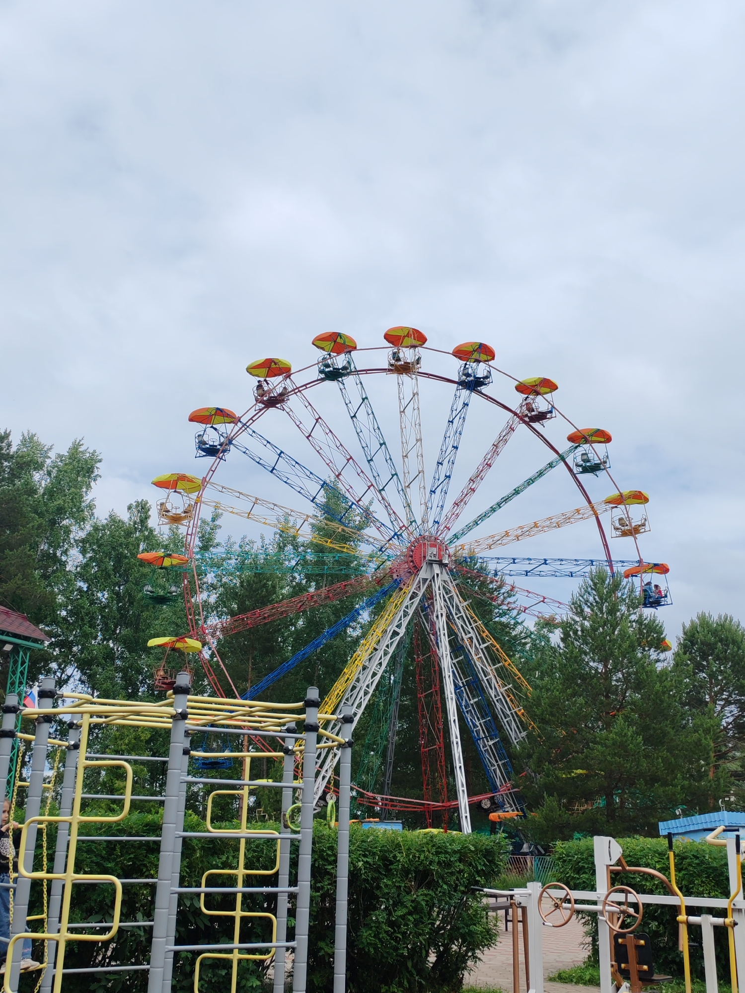 The image shows a colorful Ferris wheel in an amusement park, likely located in Chaikovsky, Russia. The Ferris wheel is the dominant feature, with its bright red, yellow, blue, and green spokes and cabins. Some of the cabins have orange and yellow umbrella-like covers. Several cabins are occupied by people, suggesting the ride is in operation.
In the foreground, there is playground equipment, including a structure with yellow bars and grey supports. To the left of the playground equipment, a young girl is visible, partially obscured. Behind the Ferris wheel and playground equipment, there are numerous green trees, some of which appear to be pine trees. The sky is overcast with grey clouds. In the lower right of the image, part of an outdoor exercise machine with wheels can be seen. The overall scene depicts a recreational area with attractions for both children and adults.