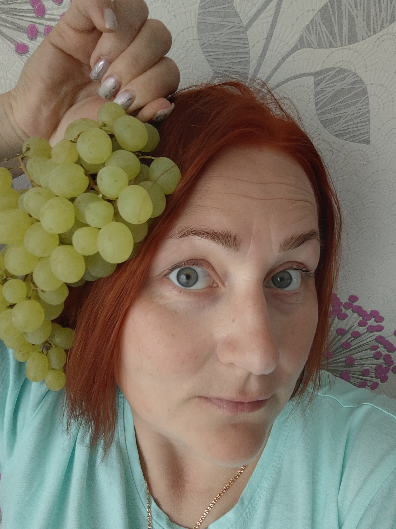 A fair-skinned woman with short, red hair and grey eyes is holding a bunch of green grapes in her left hand. She wears a light blue t-shirt and a gold chain is visible around her neck. Her fingernails are painted with a swirling silver pattern. The background appears to be a wall with a pattern of grey leaves and stylized pink floral designs. The location is Markovskii, Russia.