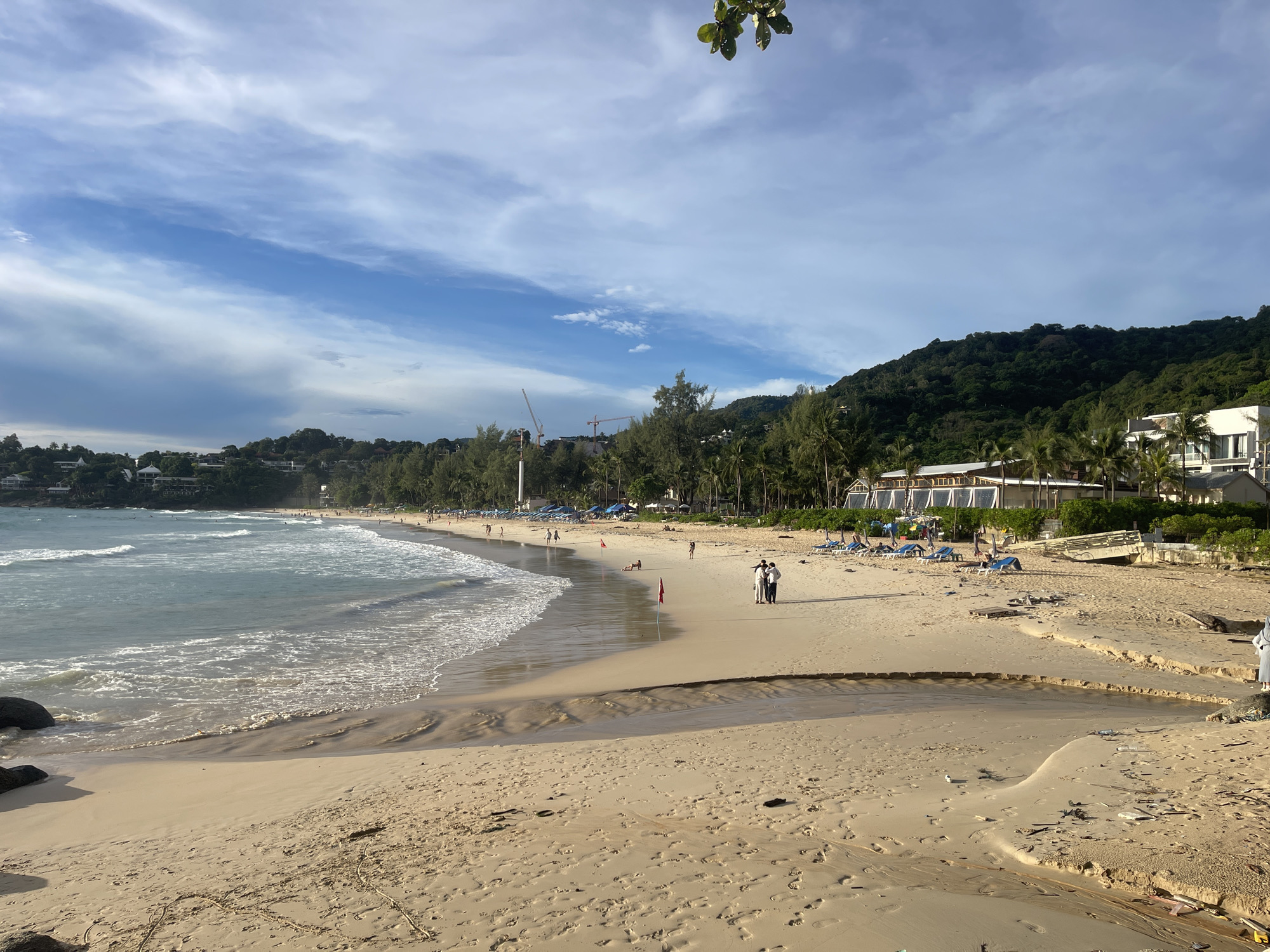 This is a wide shot of Karon Beach, Thailand, on a bright, slightly overcast day. The foreground is dominated by a sandy beach with numerous footprints. A small, sandy channel, possibly from recent rainfall, cuts across the sand. In the mid-ground, the ocean meets the shore with gentle waves. A few people are visible walking on the beach and sunbathing on loungers under umbrellas. Further back, palm trees and lush green hills form the backdrop. Buildings, some appearing to be hotels or resorts, are nestled among the trees on the hillsides. Construction cranes are visible in the distance, suggesting ongoing development. A few scattered pieces of debris, including plastic bottles and wood, are present on the sand.