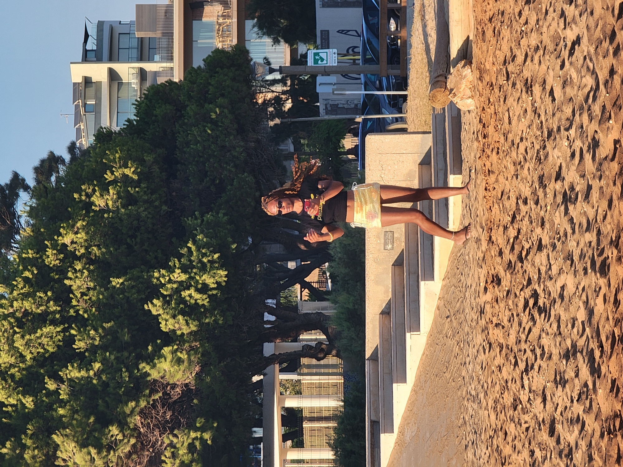A young Black woman with braided hair and sunglasses is standing on a sandy beach in Quarteira, Portugal. She is wearing a black crop top and a short, sparkly yellow skirt. She is holding a red object in her right hand and her left hand is raised. She is looking to her left. Behind her, there are stairs leading up to a building with large windows. In the background, there is a large green tree and a modern building with balconies. The sun is shining, casting long shadows on the sand. Cars are parked in the background behind a wooden fence.