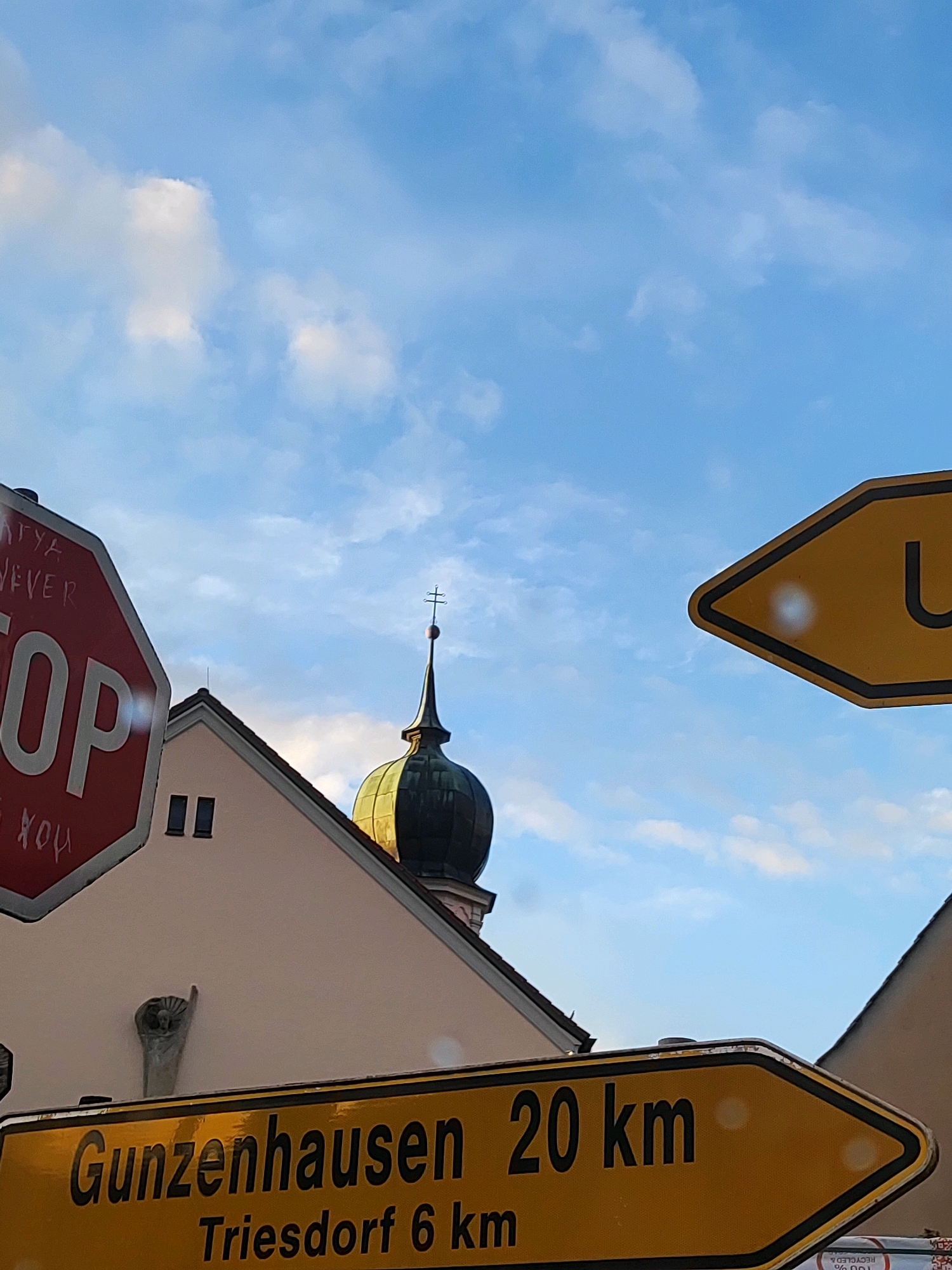 The image shows a street sign in Burgoberbach, Germany, pointing towards Gunzenhausen (20 km) and Triesdorf (6 km). A red stop sign is visible on the left, with graffiti on it. In the background, a church steeple with a cross can be seen against a blue sky with clouds. The overall scene depicts a village or town setting.