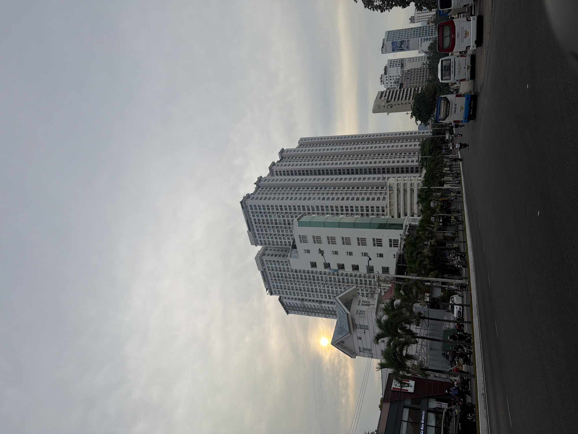 The image shows a street view in the Bac Nha Trang Ward of Vietnam. A high-rise building complex dominates the scene, with multiple towers extending upwards. A road runs along the right side of the frame, with parked buses and moving motorcycles. Along the roadside, palm trees are interspersed with parked motorcycles. A building with "Ga Kan Kentucky" signage is visible at the bottom of the frame. The sky is overcast with the sun partially obscured by clouds.