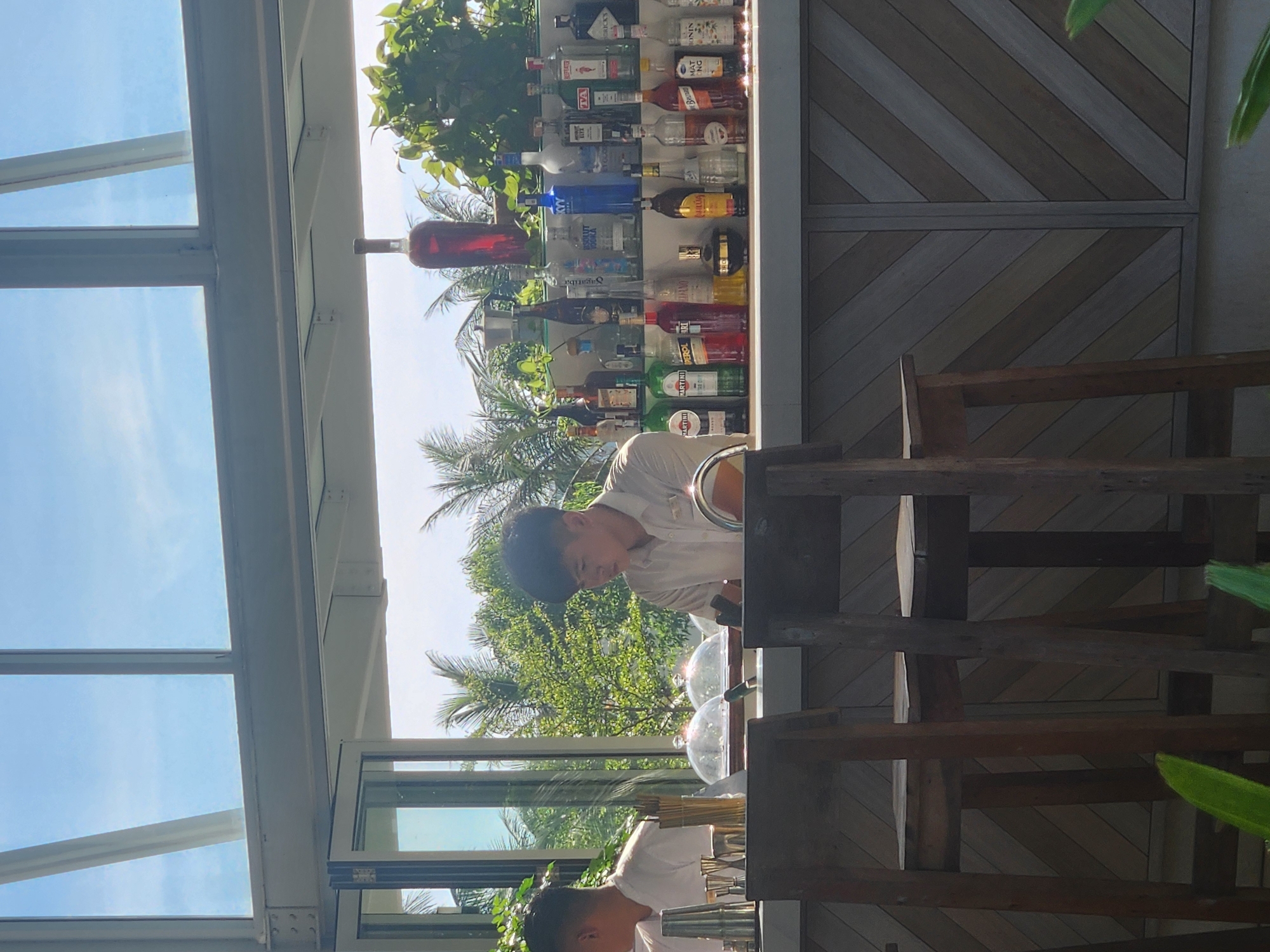 This is a medium-angle, eye-level shot of a bar area, likely at a resort or restaurant in Da Nang, Vietnam, on a bright, sunny day. The background features lush green palm trees and other foliage against a clear blue sky, suggesting an outdoor or semi-outdoor setting with a tropical ambiance.
In the foreground, two bar stools with wooden legs and seats are visible, positioned in front of a bar counter. The bar counter itself is constructed of light-colored wood arranged in a diagonal pattern.
Behind the counter, two young Asian men, presumably bartenders, are at work. The bartender on the right, wearing a light-colored collared shirt, is focused on something on the counter, possibly preparing a drink. The bartender on the left, also in a white collared shirt, is partially visible, facing away from the camera and towards the bar.
The bar top is lined with a variety of bottles, predominantly alcoholic beverages, suggesting a well-stocked bar. Brands like Martini, Aperol, and what appears to be Absolut vodka are identifiable. There are also cocktail shakers, jiggers, and other bar tools laid out. Two glass dome covers are placed on the bar, which might be protecting food items or ingredients. A tall, dark bottle with a reddish liquid, possibly a spirit like brandy or rum, stands out on a shelf behind the bar. A large red bottle of what looks like some sort of liquor or mixer is positioned prominently at the top of the shelf, catching the light.
The overall impression is one of a relaxed, upscale bar environment, likely situated in a tourist-friendly location in Da Nang, given the tropical vegetation and the appearance of the establishment. The lighting suggests it's daytime, possibly late morning or afternoon, with natural light illuminating the scene.