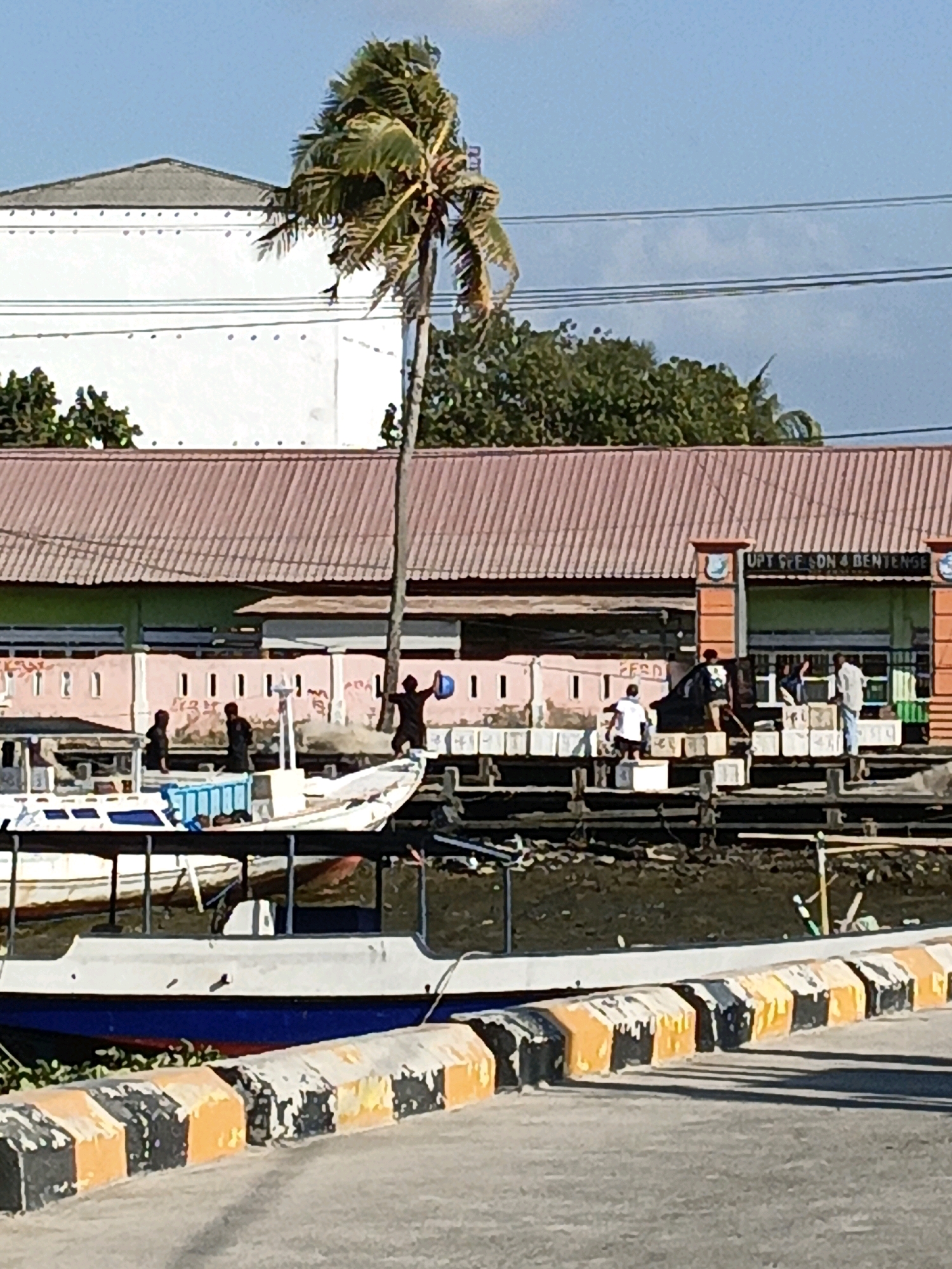 In Bulukumba, Indonesia, on a sunny day, a coastal scene unfolds with several boats moored near a building. A prominent palm tree stands tall in the foreground, its fronds rustling in the breeze. The building, with a reddish-brown corrugated roof, appears to be a public facility, possibly a government office or a terminal, as indicated by the partial signage "UPT ... SON 4 BENTENGE" visible above the entrance. Several individuals are engaged in activities on a wooden pier adjacent to the building. One person, silhouetted against the bright sky, holds a blue bucket. Others are standing or walking, suggesting various tasks or preparations related to the boats or the pier. The water reflects the clear blue sky, and the scene is bordered by a road with a distinctive black and yellow striped barrier in the lower portion of the image.